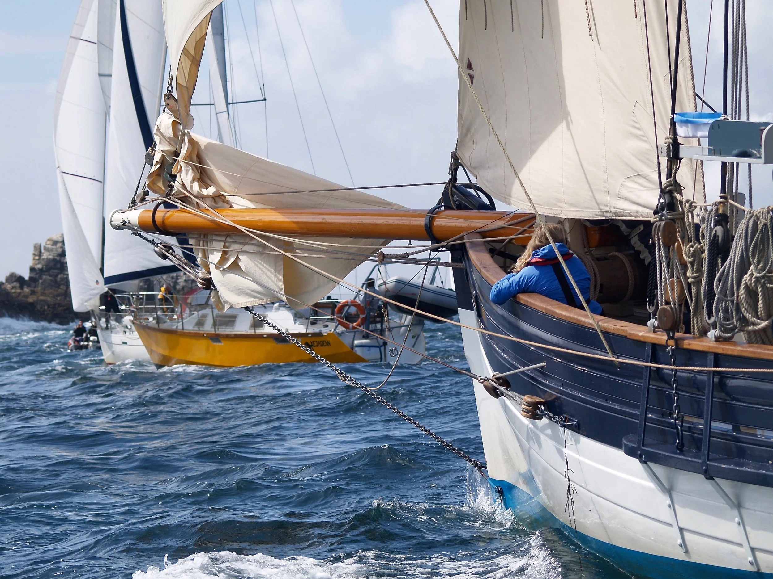 Voiliers en mer, vue de près sur la coque et les voiles, avec des personnes à bord, en navigation par temps ensoleillé avec ciel partiellement nuageux.