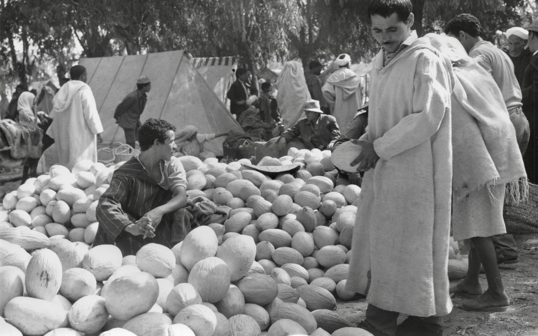 Vendeurs et acheteurs dans un marché en plein air, avec des melons et autres fruits sur les tables, scène en noir et blanc.