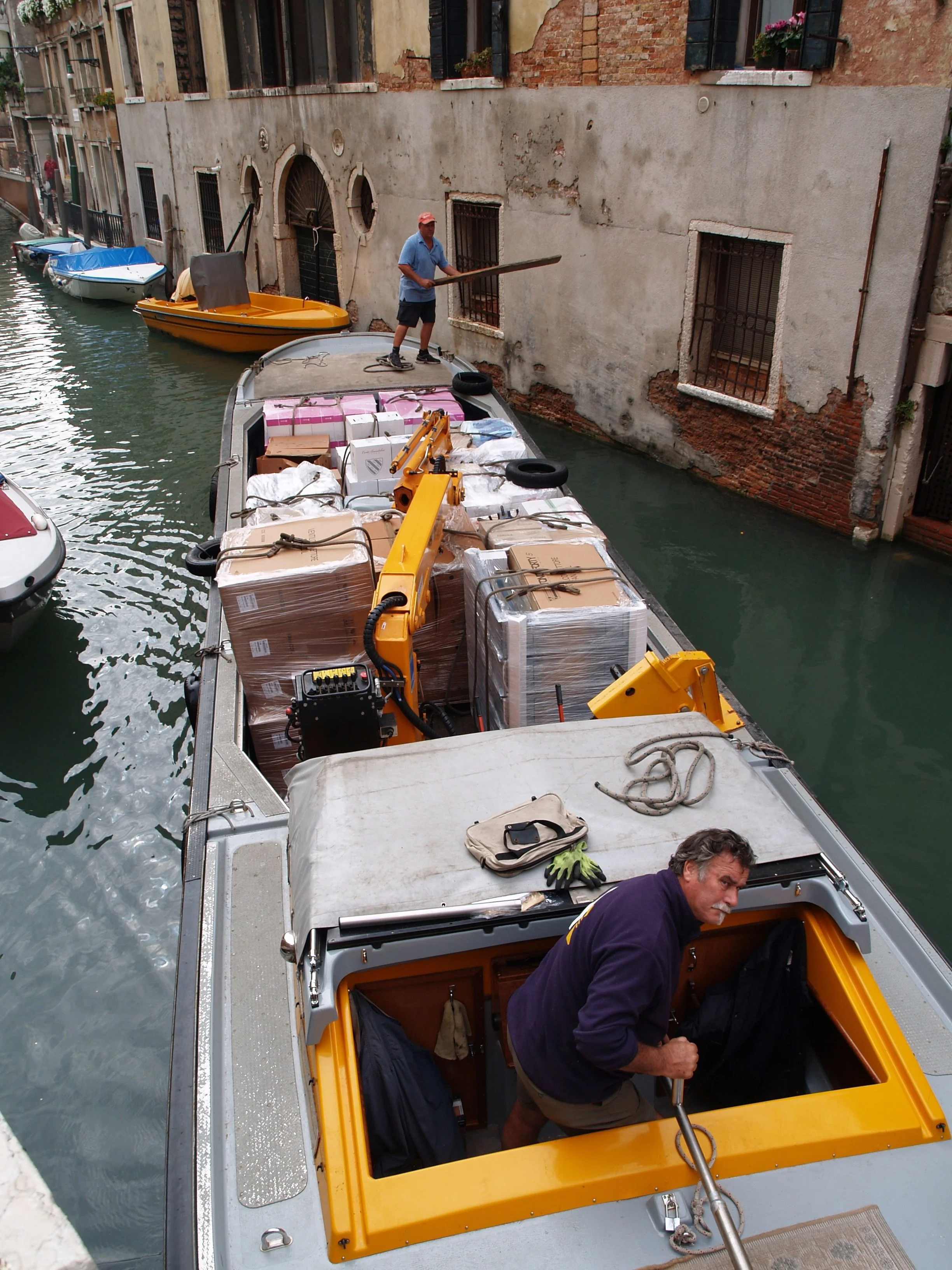 Une scène dans un canal de Venise où des personnes chargent ou déchargent un petit bateau, avec des bâtiments anciens en fond.