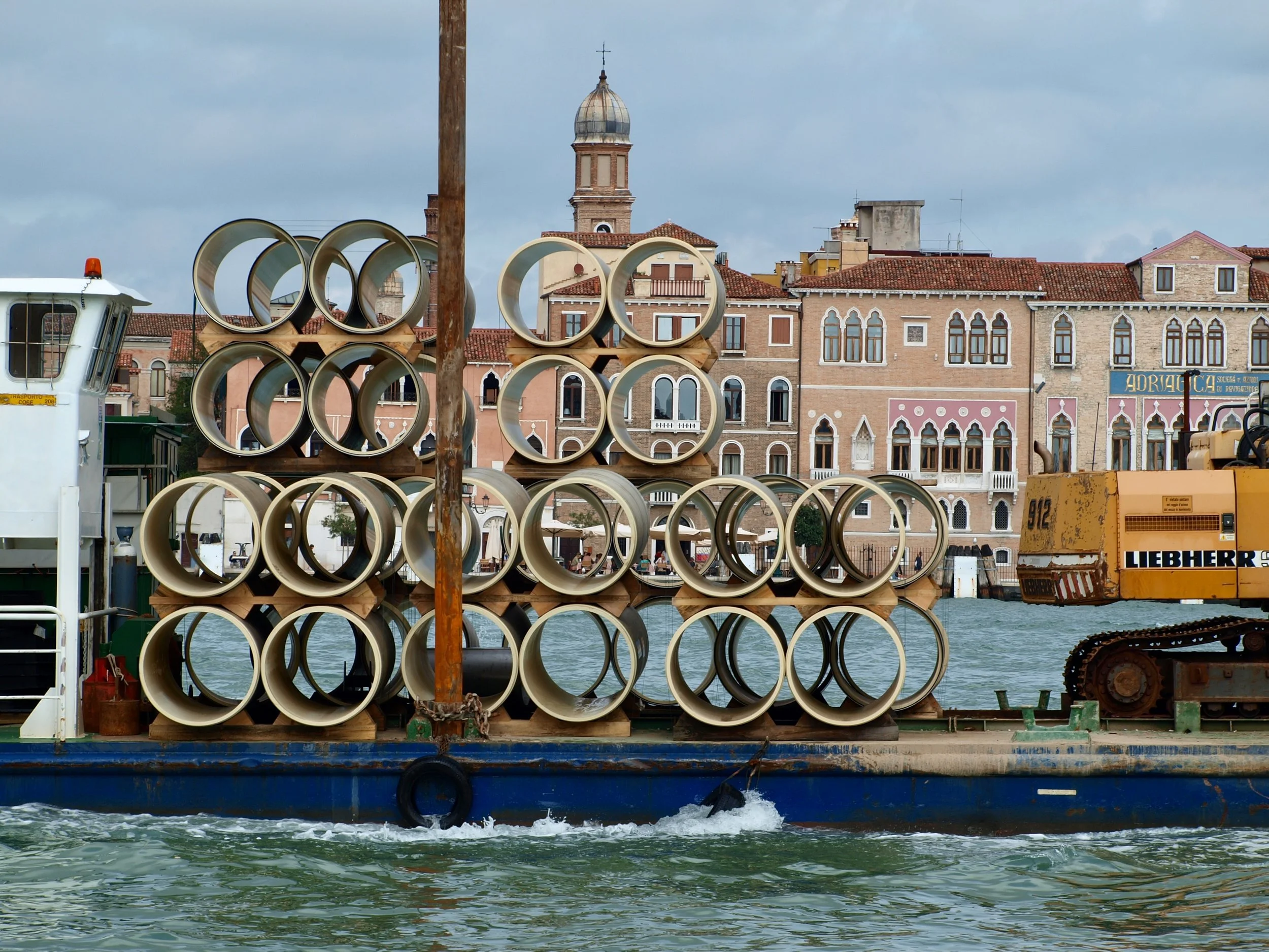 Une scène urbaine vue d'une embarcation sur l'eau avec une pile de tuyaux de construction en premier plan, des bâtiments anciens avec des fenêtres en arche derrière et un ciel nuageux.