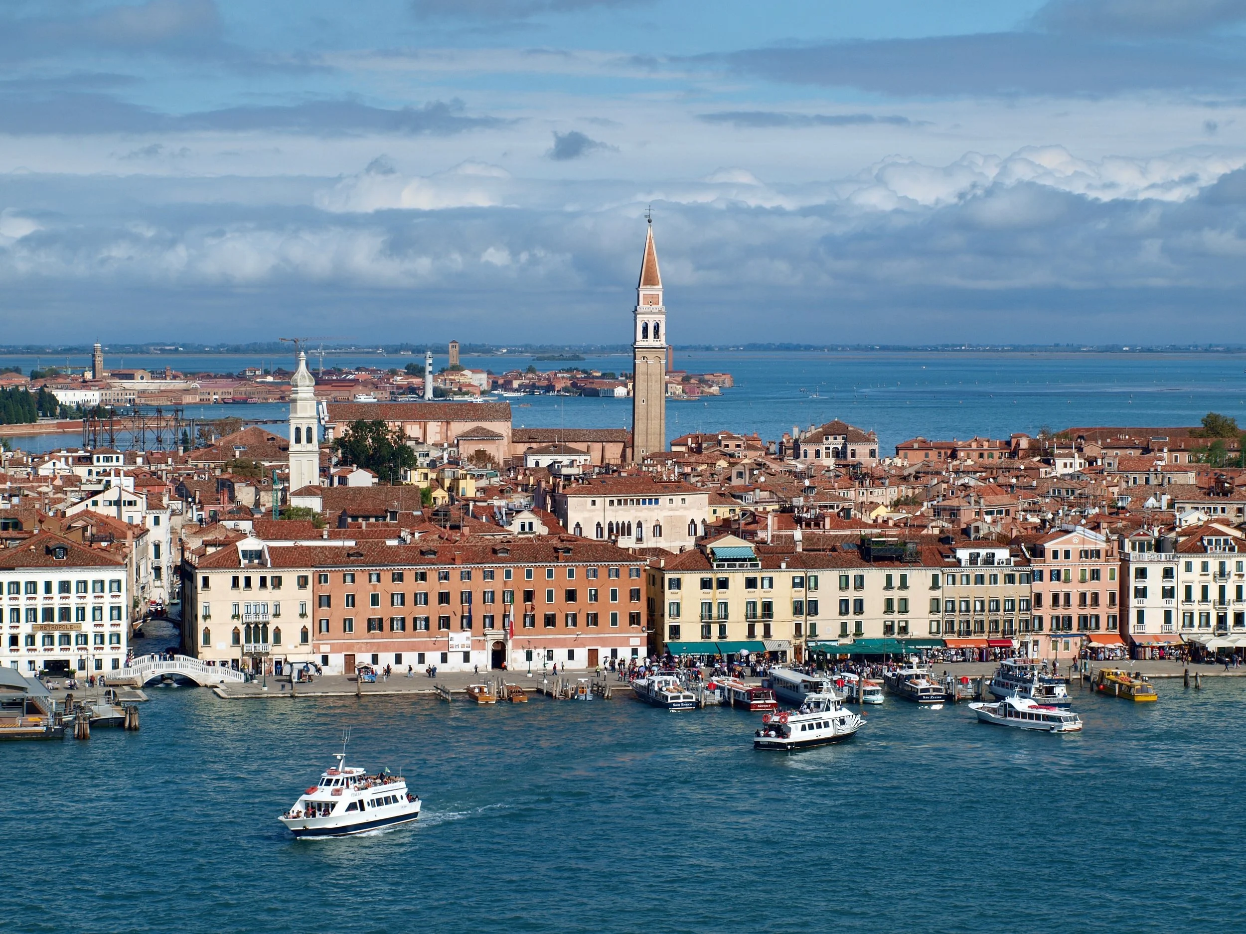 Vue d'ensemble d'une ville située au bord d'une grande étendue d'eau, avec un port, des bateaux, des bâtiments aux toits rouges, et une tour avec un clocheton, sous un ciel partiellement nuageux.