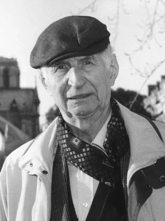 Un homme âgé portant une casquette noire et un manteau clair, avec un foulard autour du cou, dans un environnement urbain en plein air.