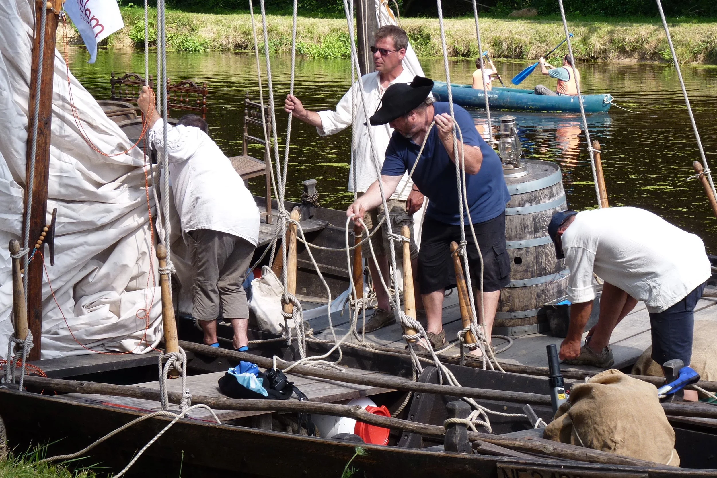 Groupe de personnes sur un bateau en bois, préparant une voile, avec un décor de rivière et de verdure en arrière-plan.