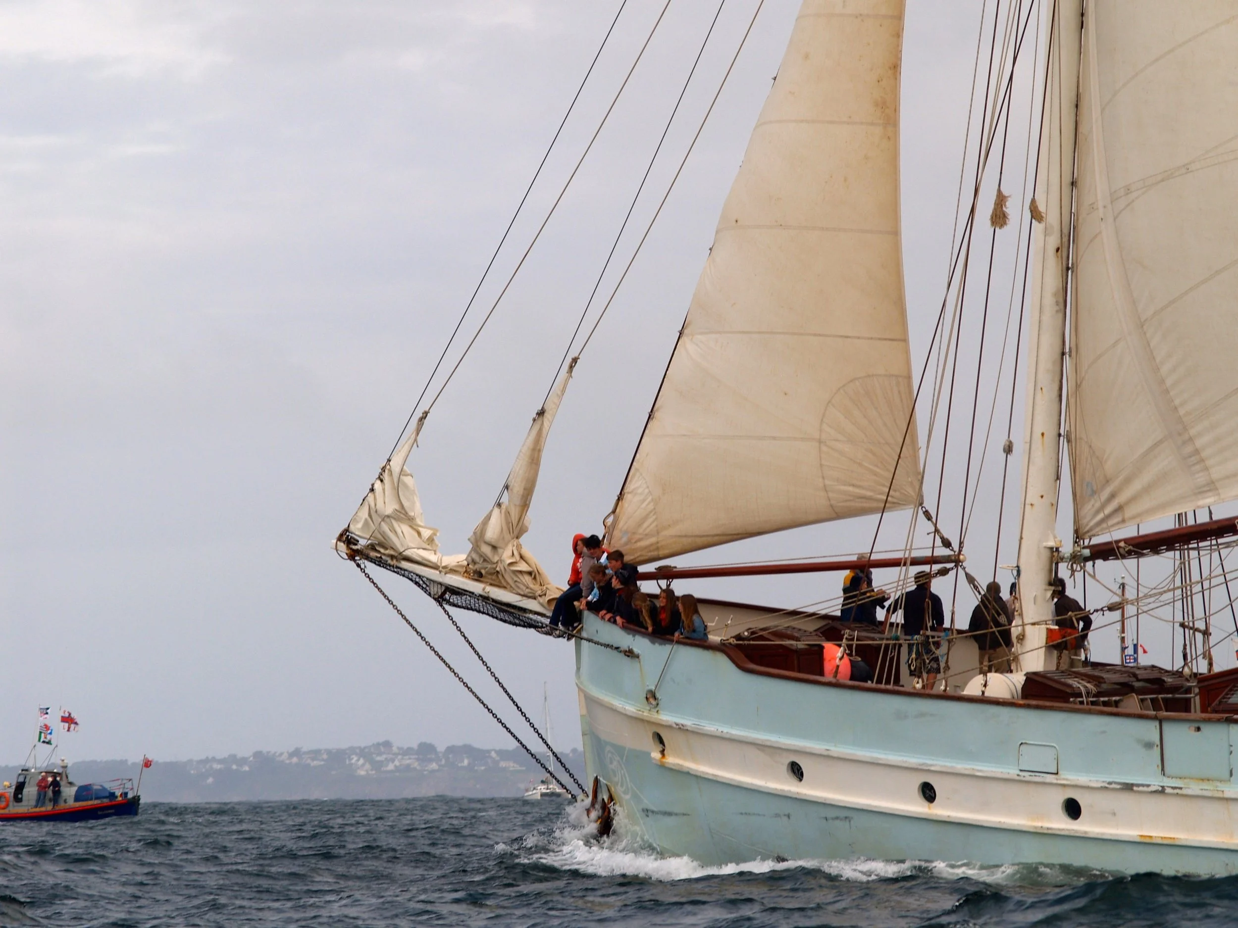 Un voilier avec plusieurs personnes à bord navigue sur la mer, avec d'autres bateaux visibles au loin.
