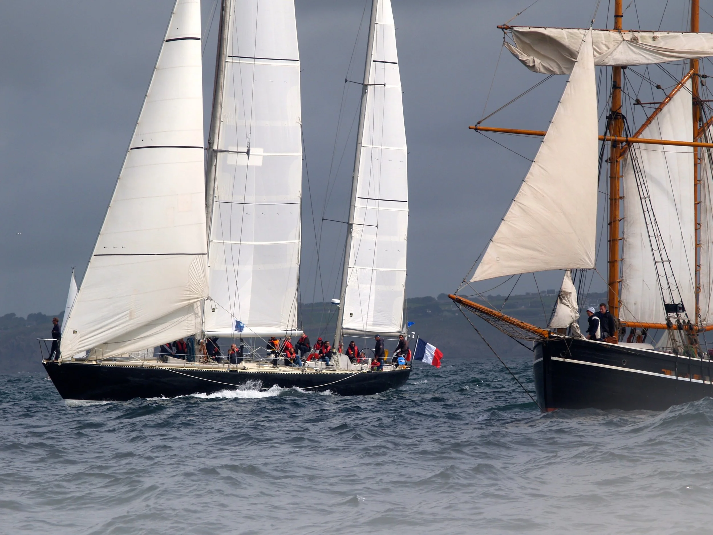 Deux voiliers de course avec leurs voiles déployées naviguent sur une mer agitée par un temps nuageux, avec plusieurs personnes à bord. L’un des voiliers est noir avec des voiles blanches, tandis que l’autre a un mât en bois et des voiles blanches.