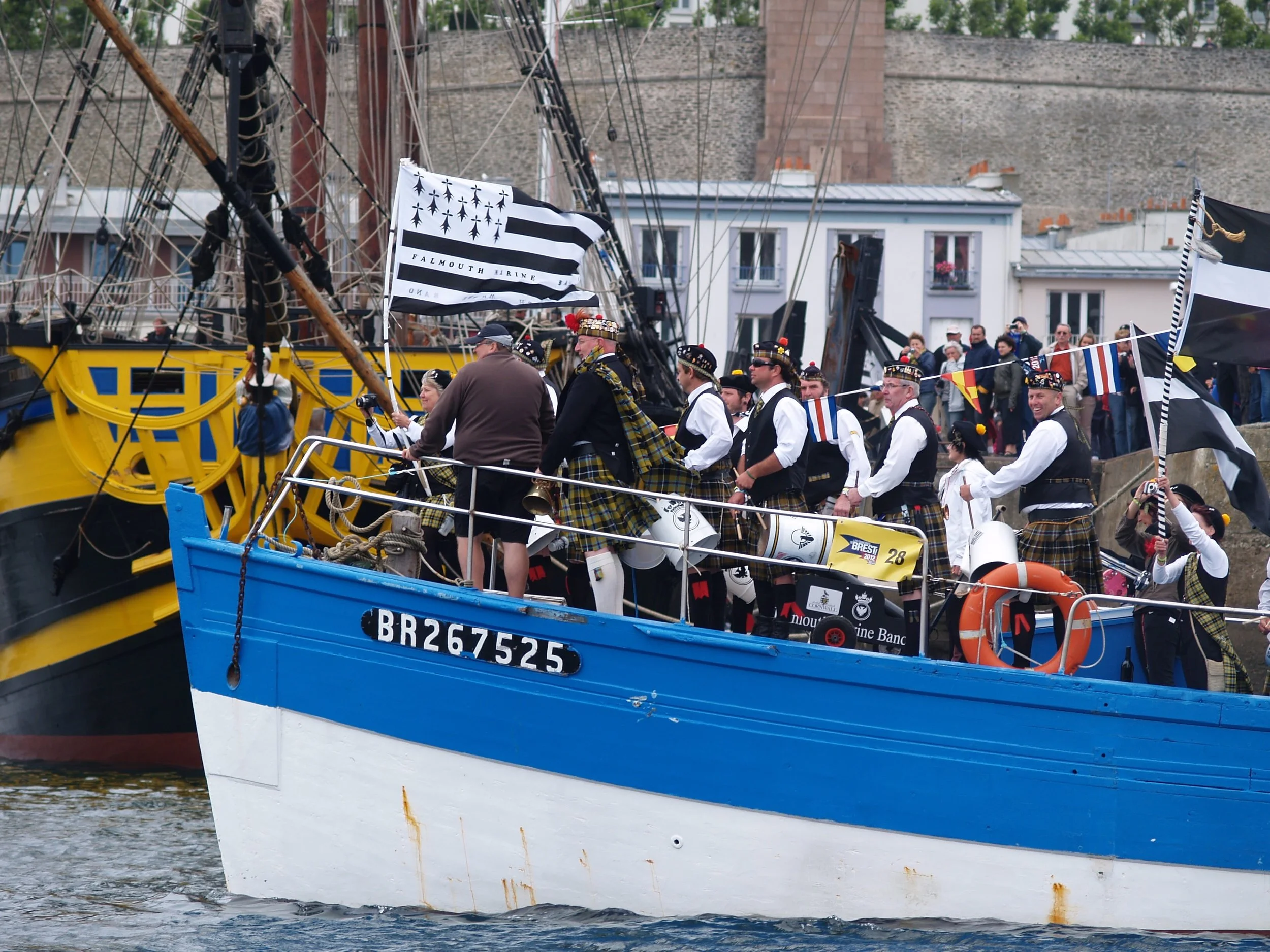 Des personnes en tenue traditionnelle écossaise festoyent et défilent à bord d’un voilier, avec des drapeaux noirs et blancs, dans un port, en arrière-plan des maisons et une digue.