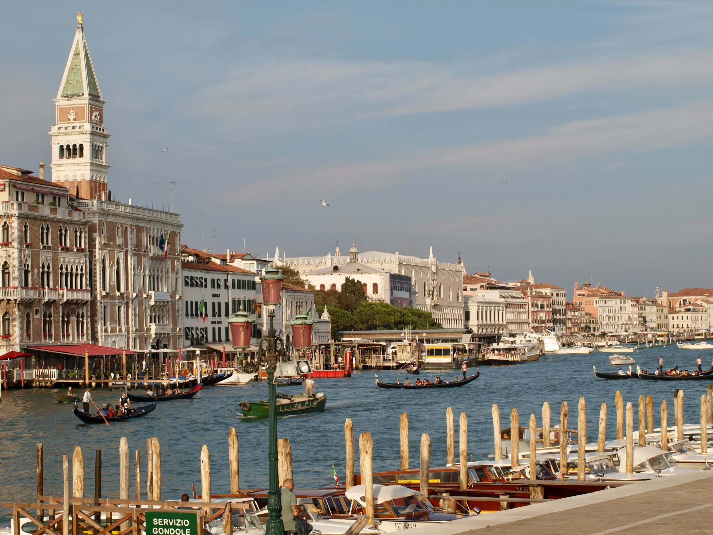 Le paysage montre la Grand Canal de Venise avec des gondoles naviguant, bordé par des bâtiments historiques et une église avec un clocher, sous un ciel partiellement nuageux.