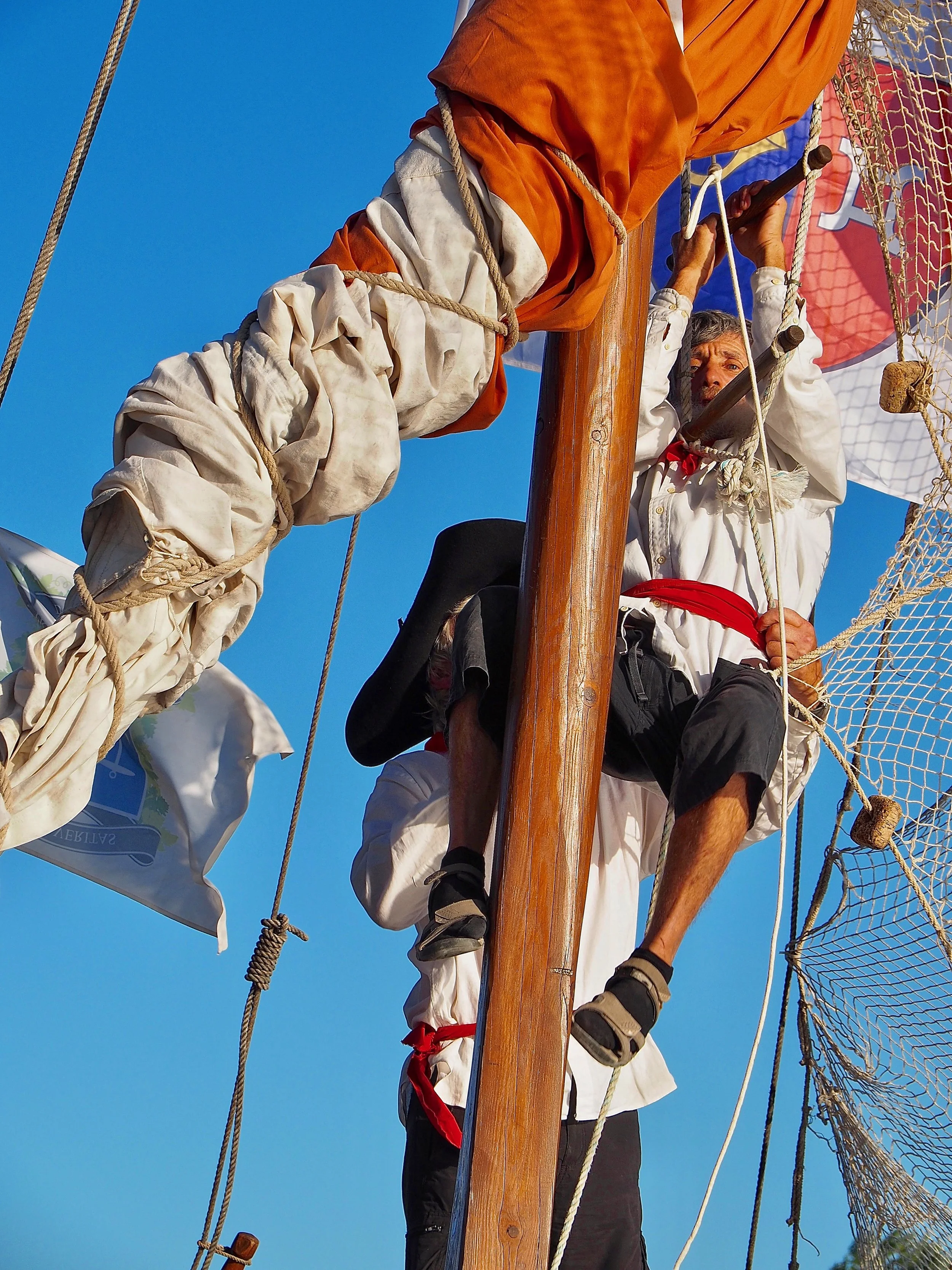 Deux hommes habillés en vêtements traditionnels blancs, avec des foulards rouges, sont sur un mât de navire, entourés de cordages et d'une voile, contre un ciel bleu, semblant être en pleine navigation ou cérémonie maritime.