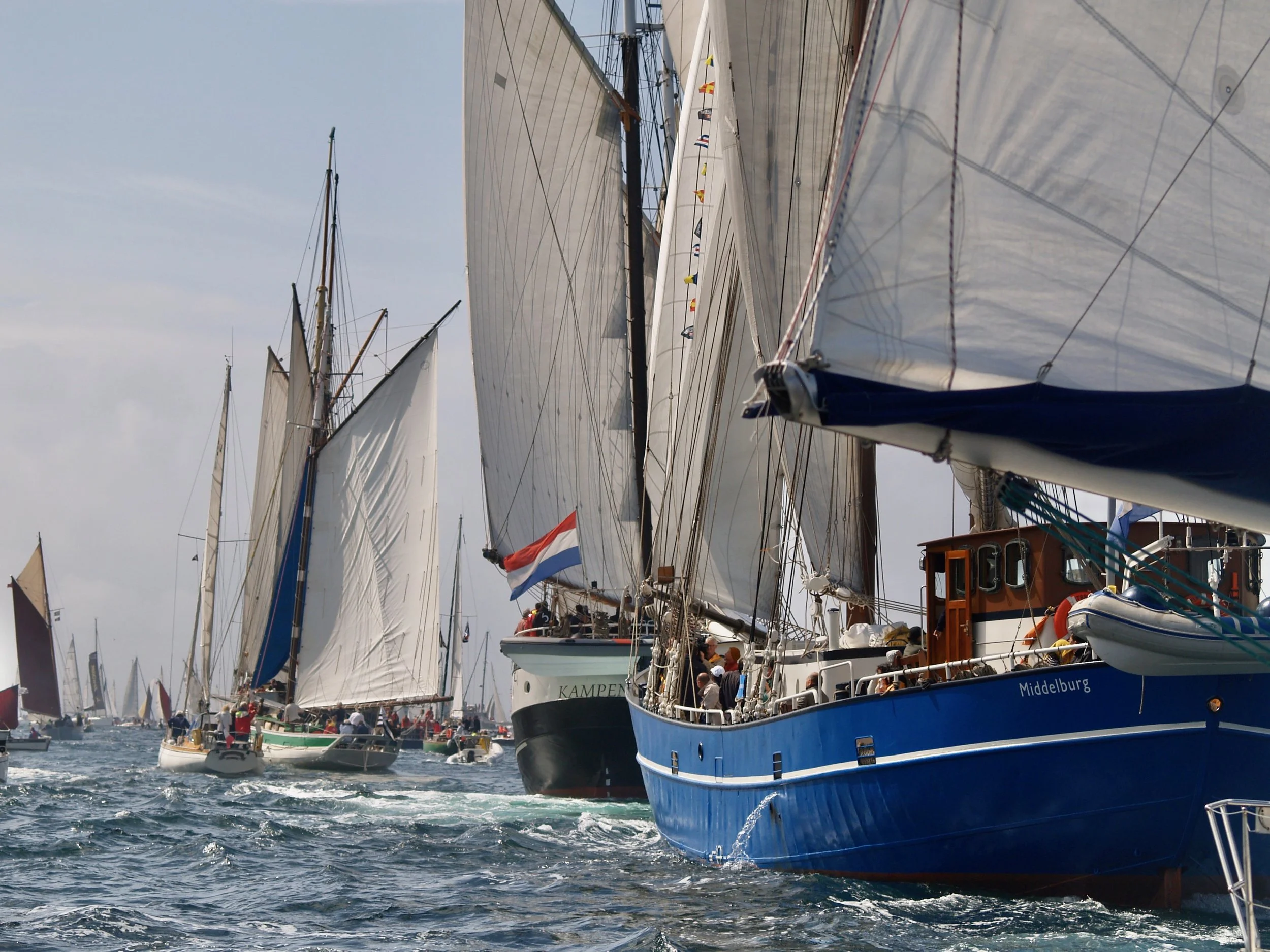 Des yachts à voile naviguent dans un port, avec beaucoup de grandes voiles blanches déployées et des équipages à bord. Certaines personnes sont visibles sur le pont du yacht bleu nommé 'Middelburg'.