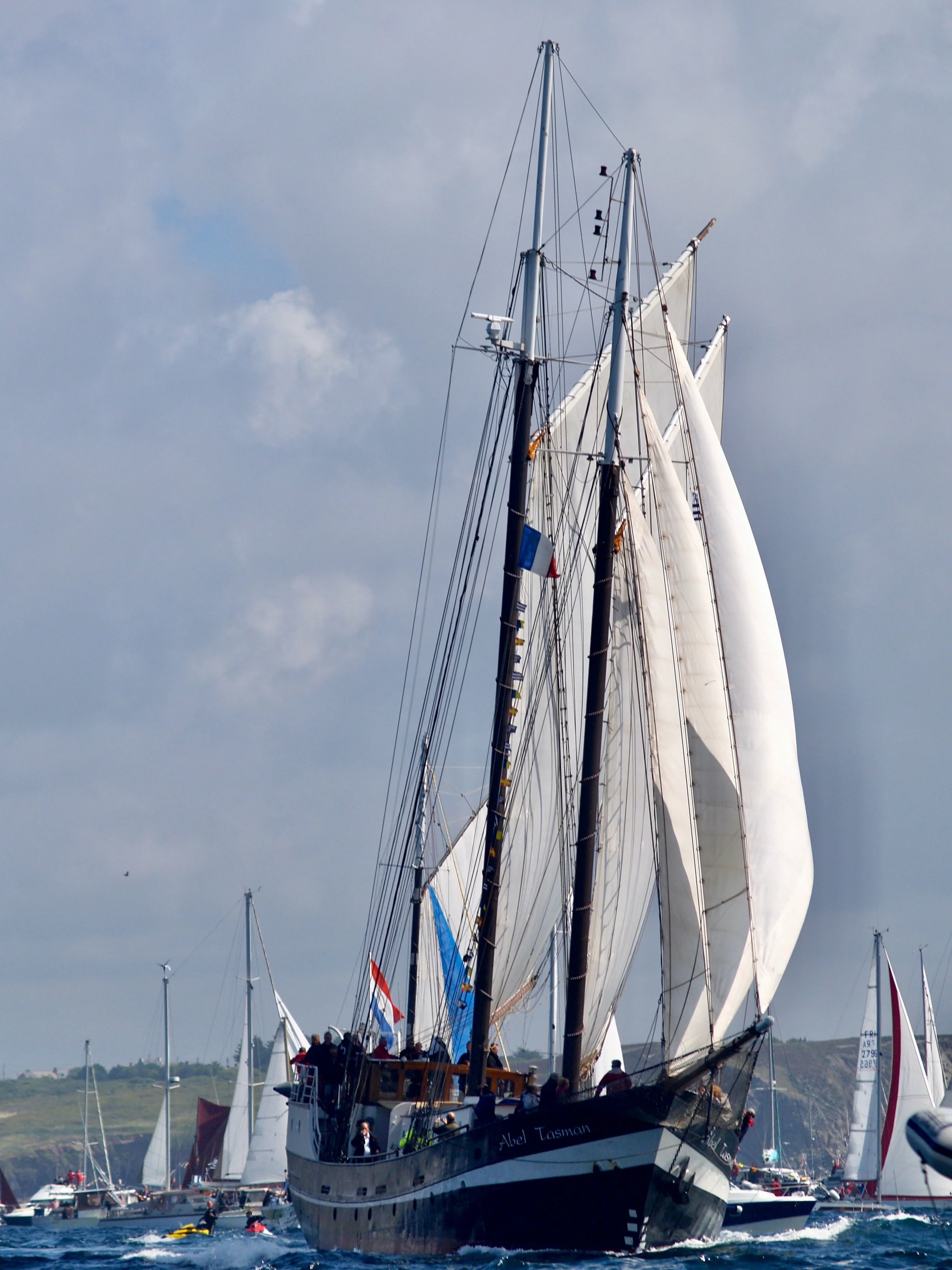 Un grand voilier avec plusieurs voiles blancs, naviguant dans un port, avec des personnes à bord et d'autres bateaux autour, sous un ciel nuageux.