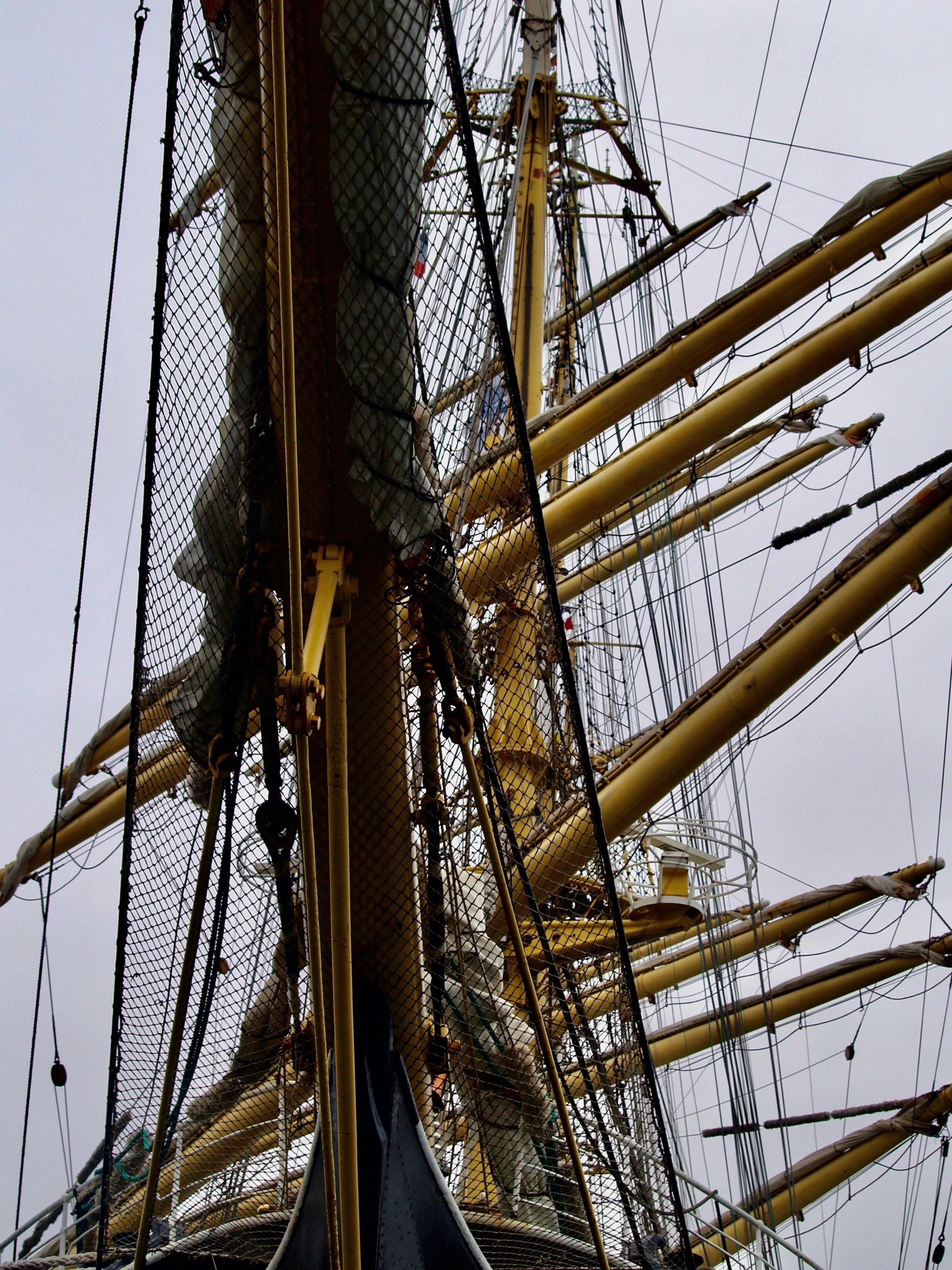 Une vue du mat et des cordages d'un grand voilier en mer, avec une plateforme d'accès visible, sous un ciel gris.
