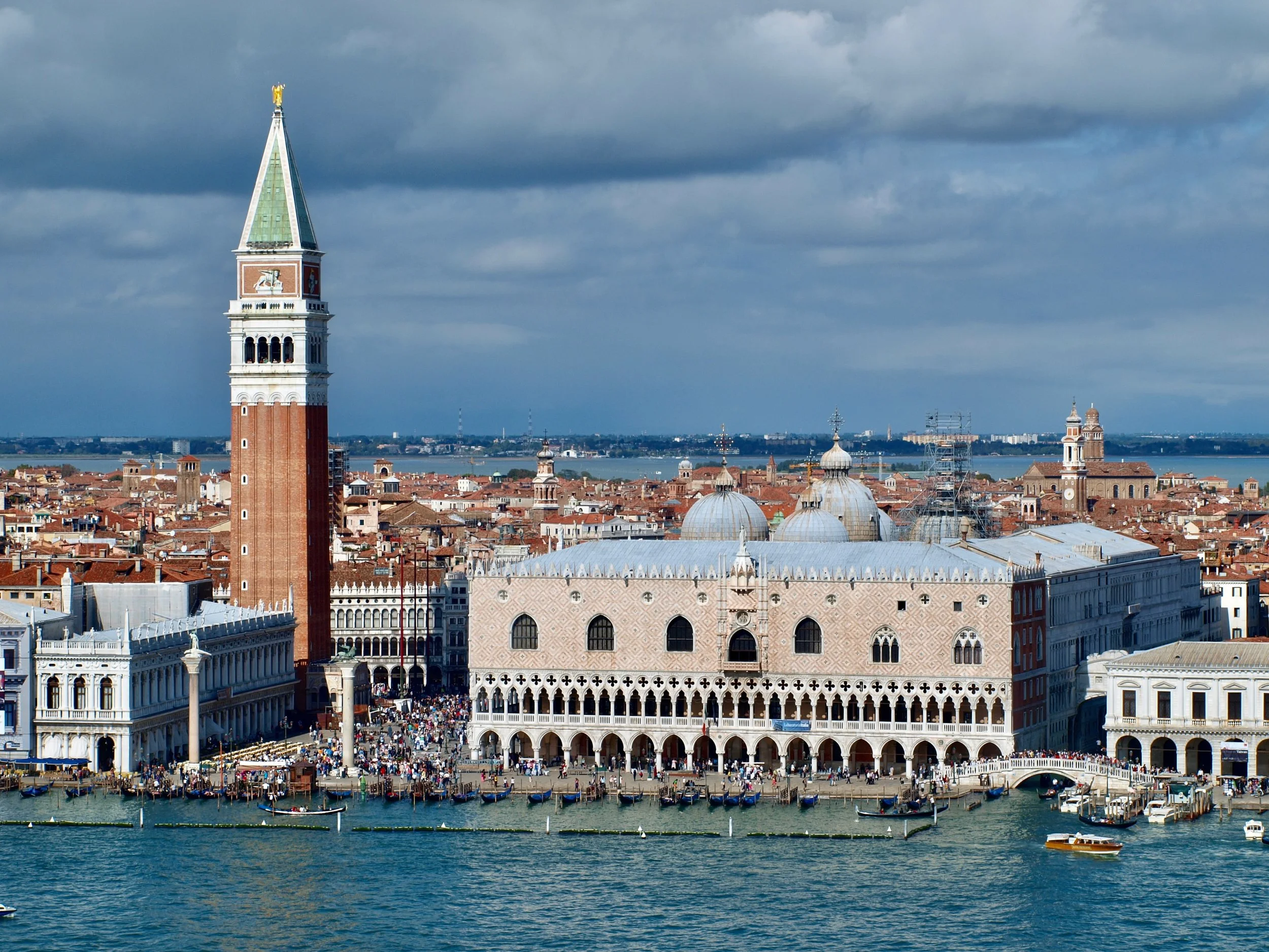 Une vue panoramique de la célèbre place Saint-Marc à Venise, avec la tour de Campanile en premier plan et les bâtiments historiques, le grand canal avec des gondoles et un ciel nuageux en arrière-plan.
