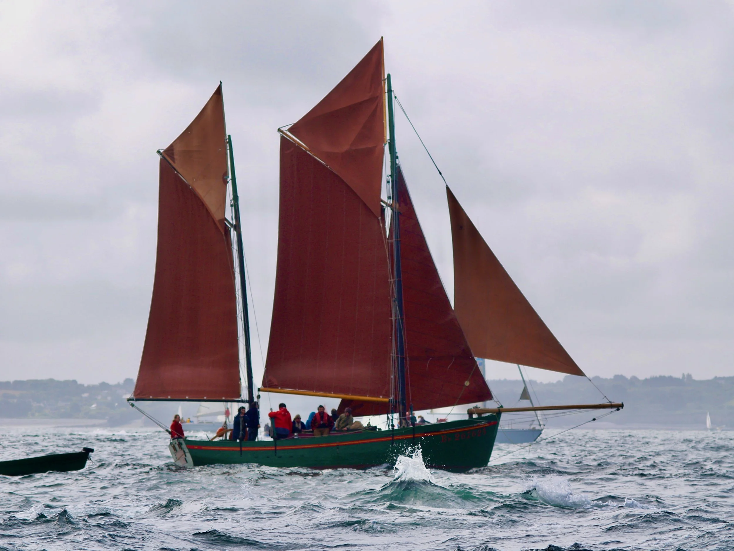Un voilier avec des voiles en tissu rouge et plusieurs personnes à bord naviguant sur l'eau, avec un ciel nuageux en arrière-plan.