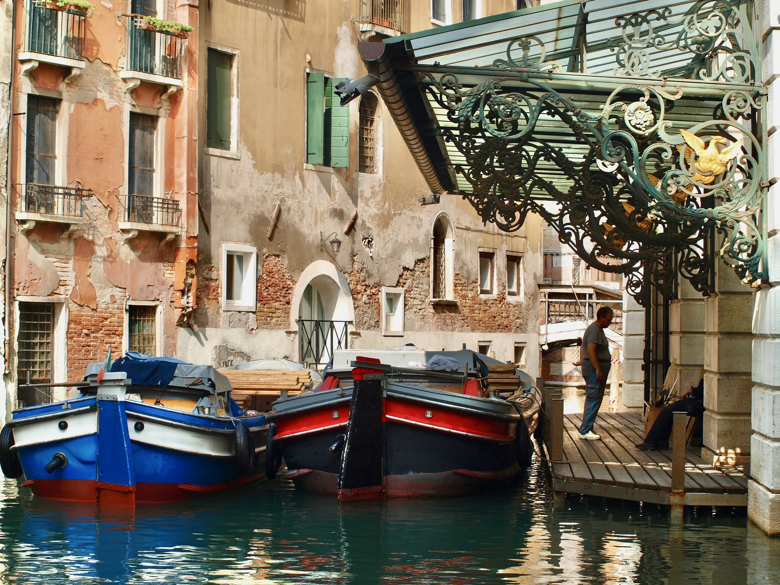 Bateaux de pêche amarrés dans une petite crique vénitienne avec bâtiments anciens en arrière-plan, quelques fenêtres avec volets verts, une façade délabrée et un balcon avec des plantes.