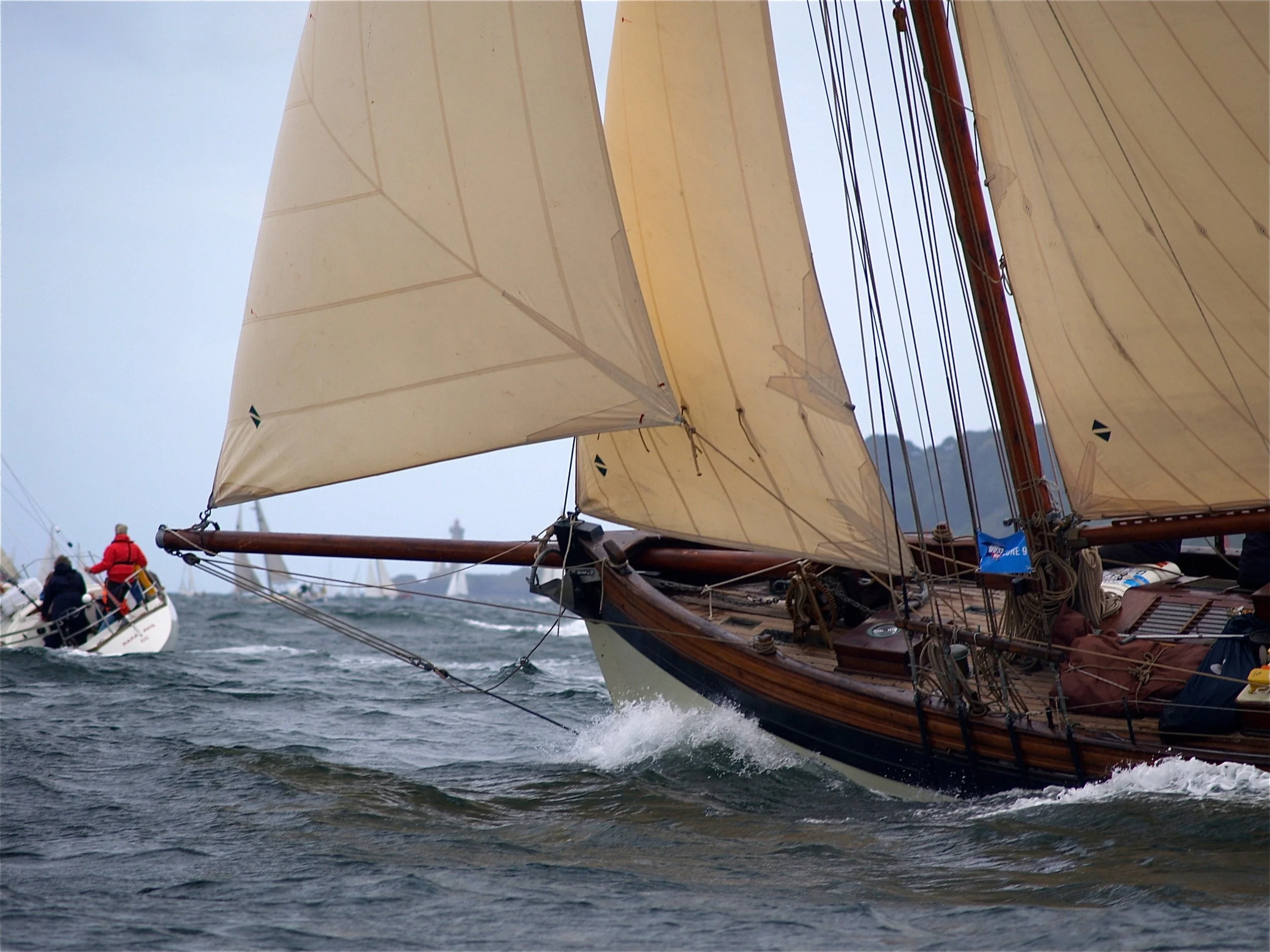 Un voilier en mer avec des voiles beige et une coque en bois, naviguant à grande vitesse, avec des personnes à bord portant des vêtements de pluie, et un autre bateau visible en arrière-plan.
