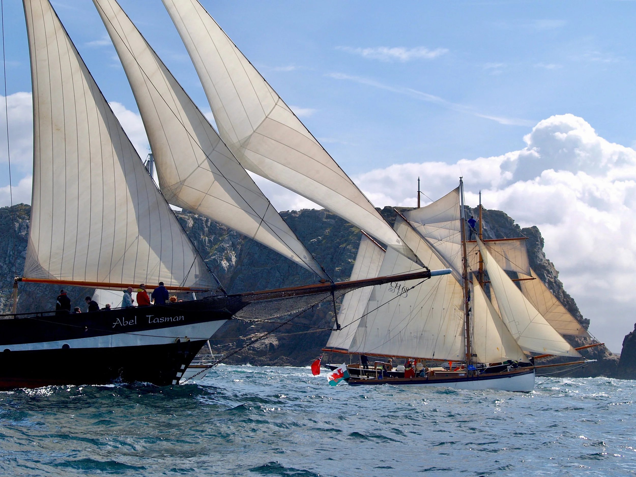 Des voiliers traditionnels avec de grandes voiles blanches naviguent en mer sous un ciel partiellement nuageux, avec une côte rocheuse en arrière-plan.