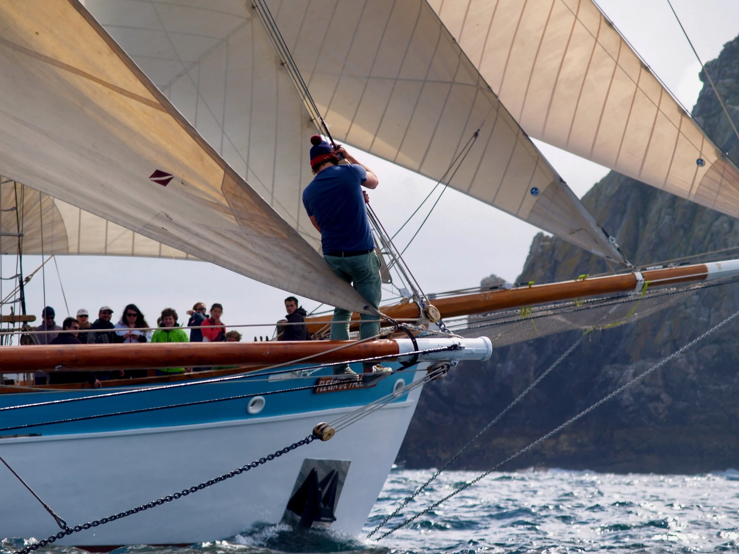 Groupe de personnes regardant une personne en train de manœuvrer un voilier avec de grandes voiles en mer, avec des montagnes en arrière-plan.