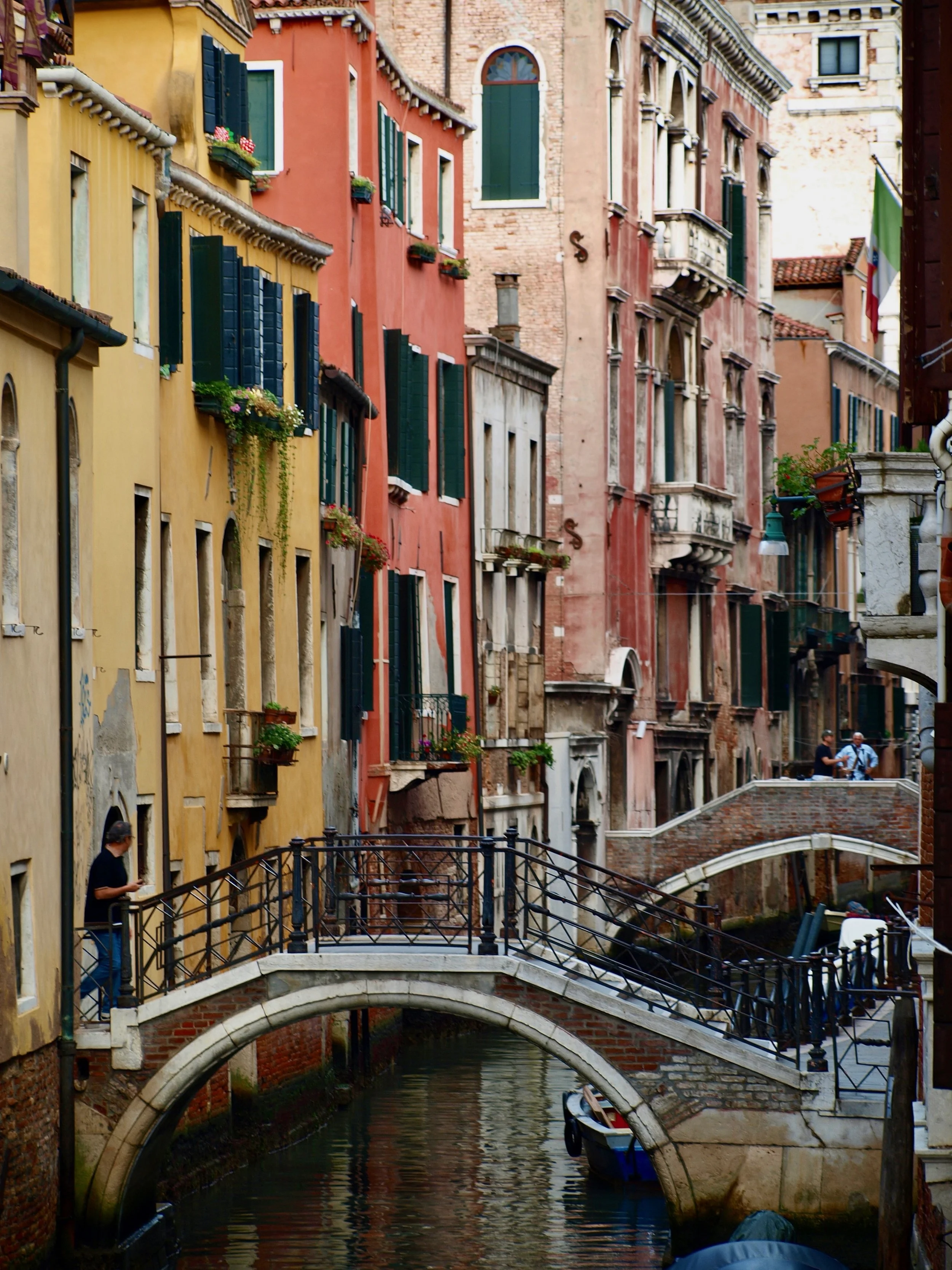 Rues colorées avec bâtiments anciens et fenêtres à volets, un petit canal avec bateau, un pont en pierre, des personnes discutant à l'arrière.