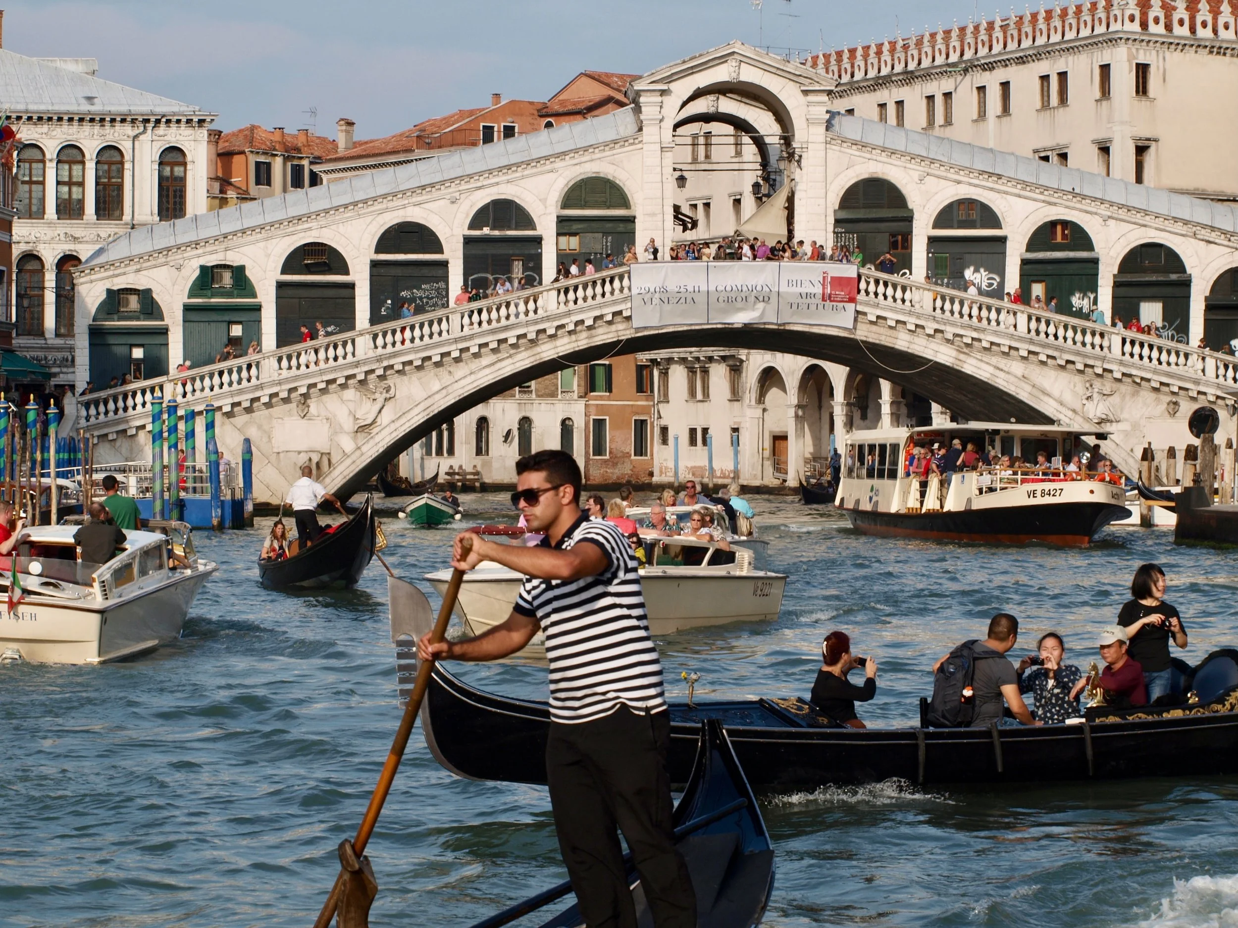 Une scène animée sur un canal à Venise avec des gondoles et un bateau de touristes. En arrière-plan, un pont en pierre avec des gens dessus, et des bâtiments historiques avec des terrasses. C'est une journée ensoleillée avec un ciel légèrement nuageu