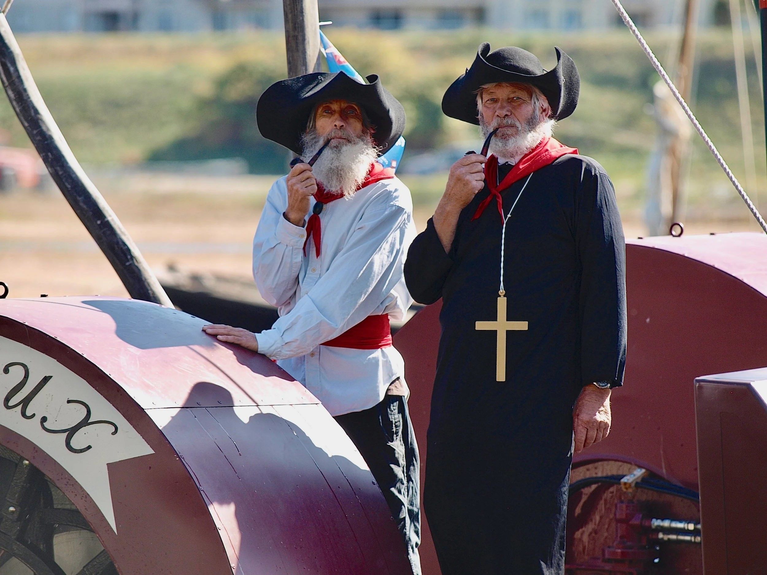 Deux hommes âgés en costumes de pirates avec de grands chapeaux noirs, barbe blanche, portant une pipe, posent à côté d'une charrette à voile dans un champ en plein air.