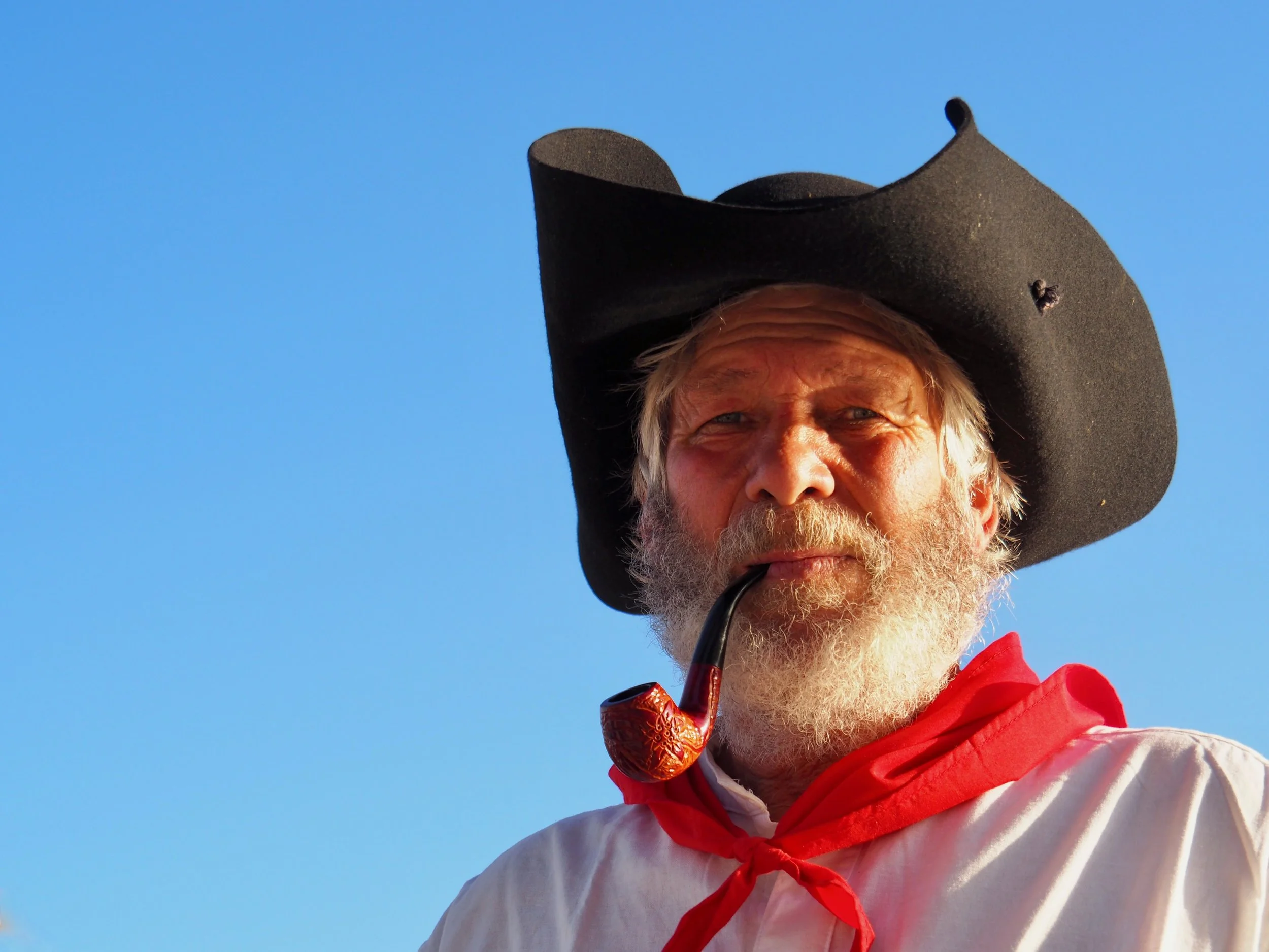 Un homme âgé portant un chapeau noir, une veste rouge et une chemise blanche, fumant une pipe en bois, avec un ciel bleu clair en arrière-plan.