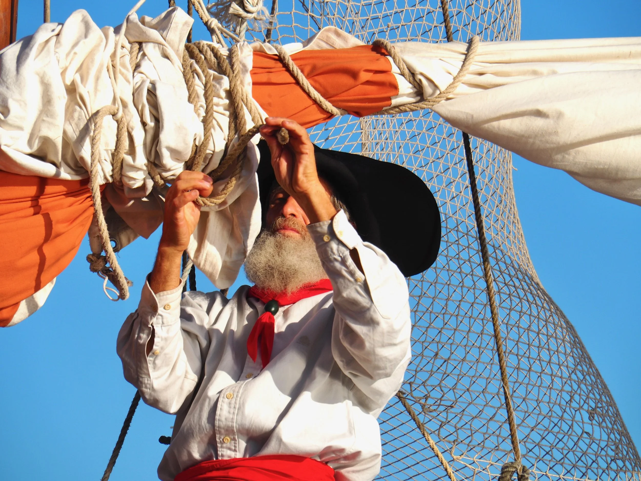 Homme âgé portant un chapeau noir, une chemise blanche, une écharpe rouge et une barbe blanche, attaché à une structure en corde dans un ciel bleu clair.