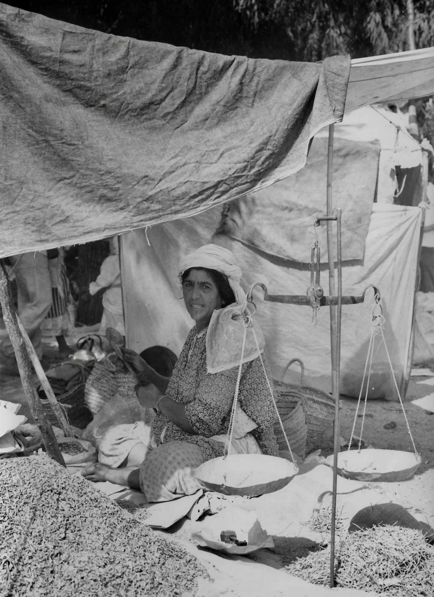Une femme assise dans un marché aux textiles avec des vêtements et des tissus, portant un turban ou un foulard, entourée de paniers et de tissus accrochés pour la vente.