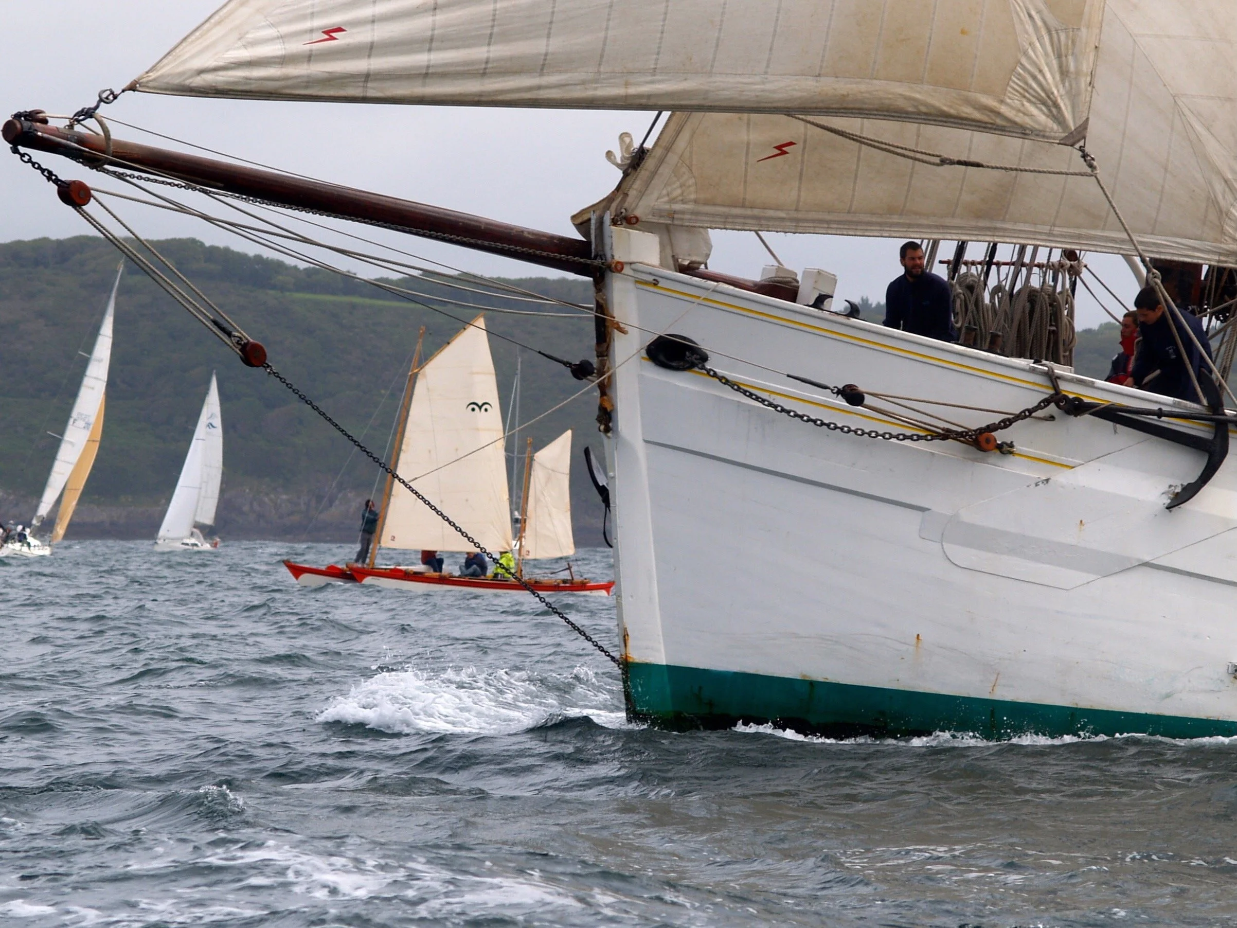 Bateau à voile naviguant en mer avec d'autres voiliers en arrière-plan, surfond de collines verdoyantes, ciel nuageux.