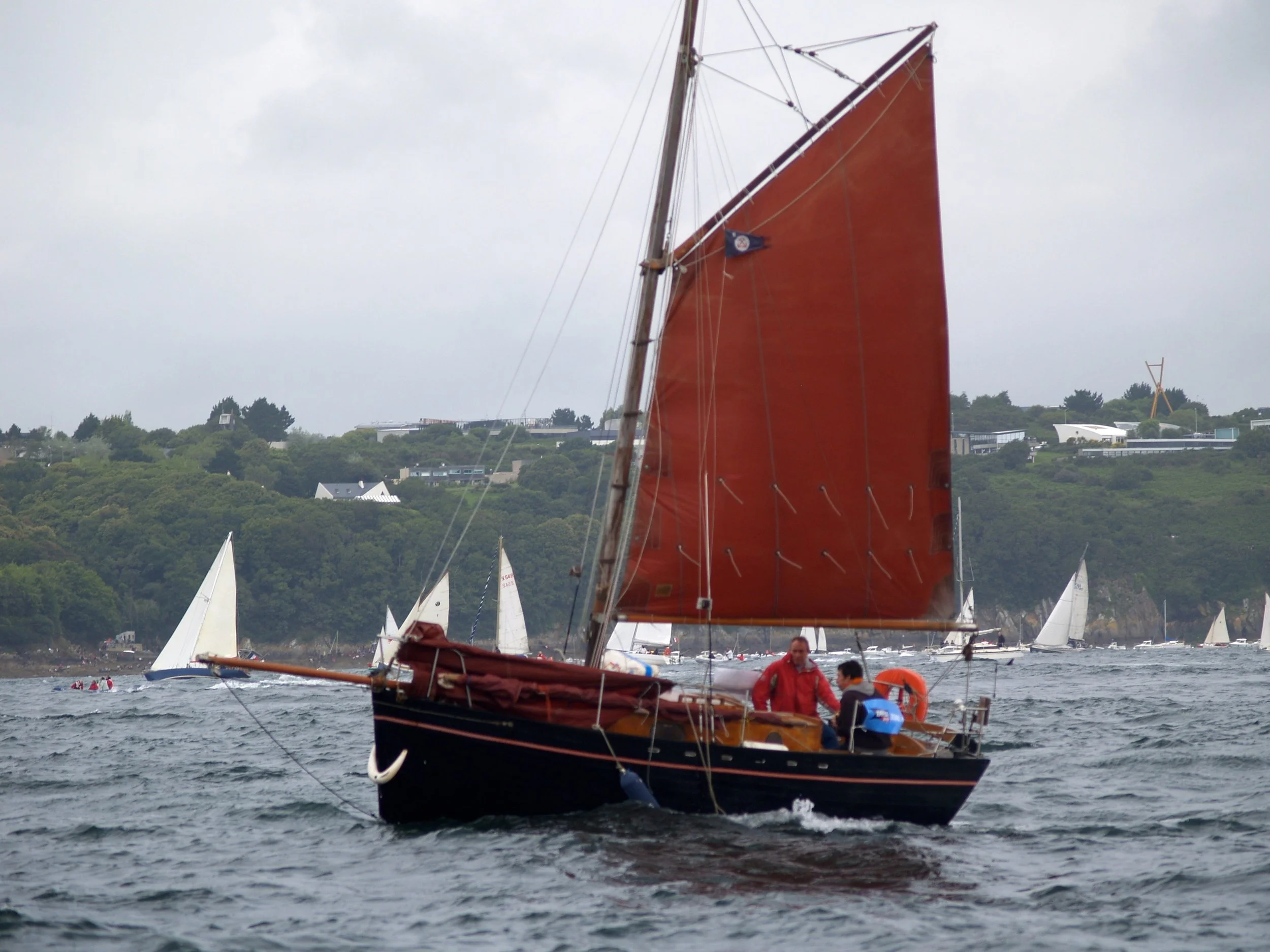 Un voilier avec une grande voile rouge navigue sur l'eau, avec plusieurs autres voiliers en arrière-plan et des collines verdoyantes en vue. Le ciel est nuageux.