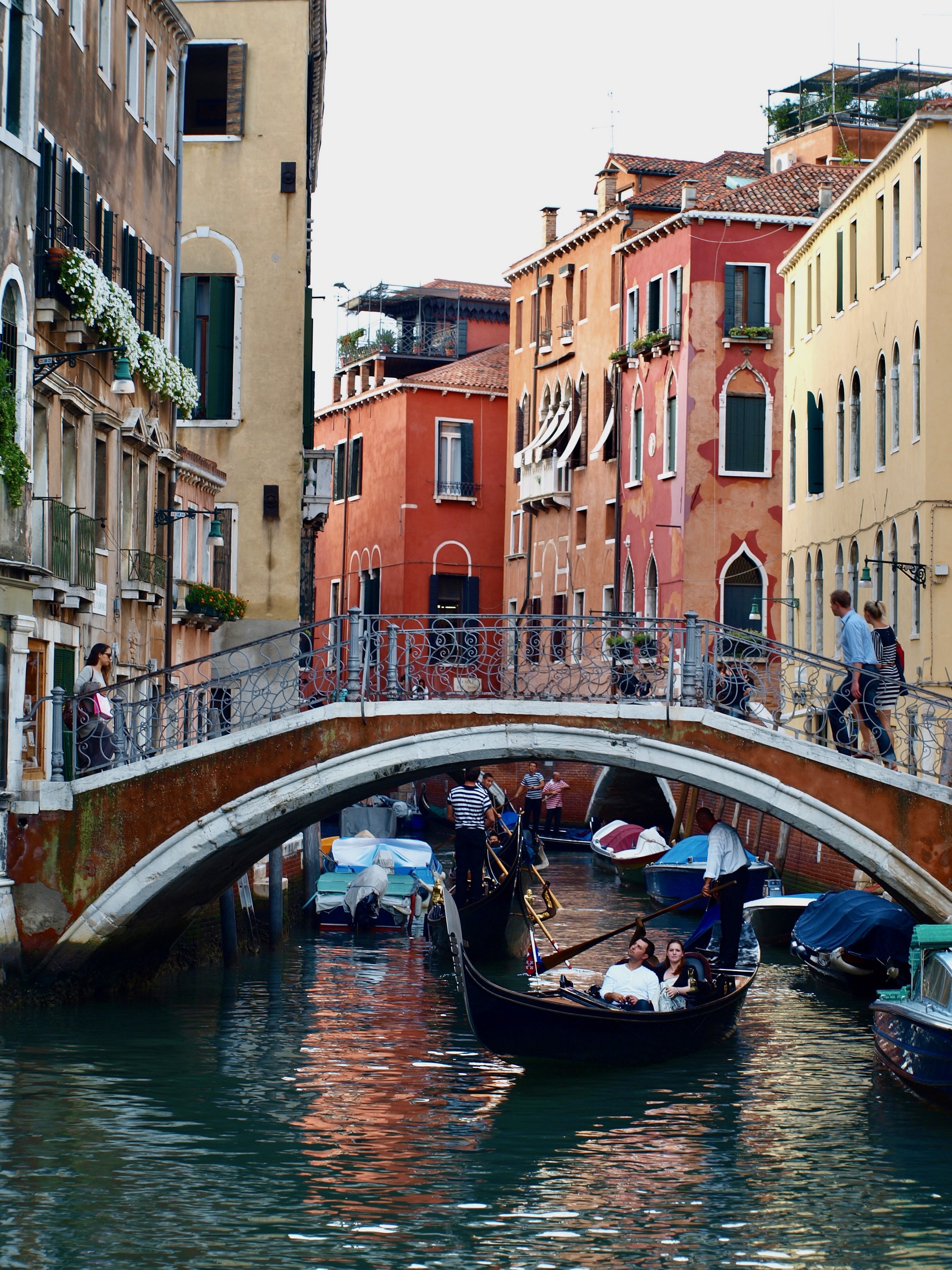 Canal à Venise avec des bâtiments colorés, un pont en pierre, et un gondolier avec des touristes à bord, en fin d'après-midi.