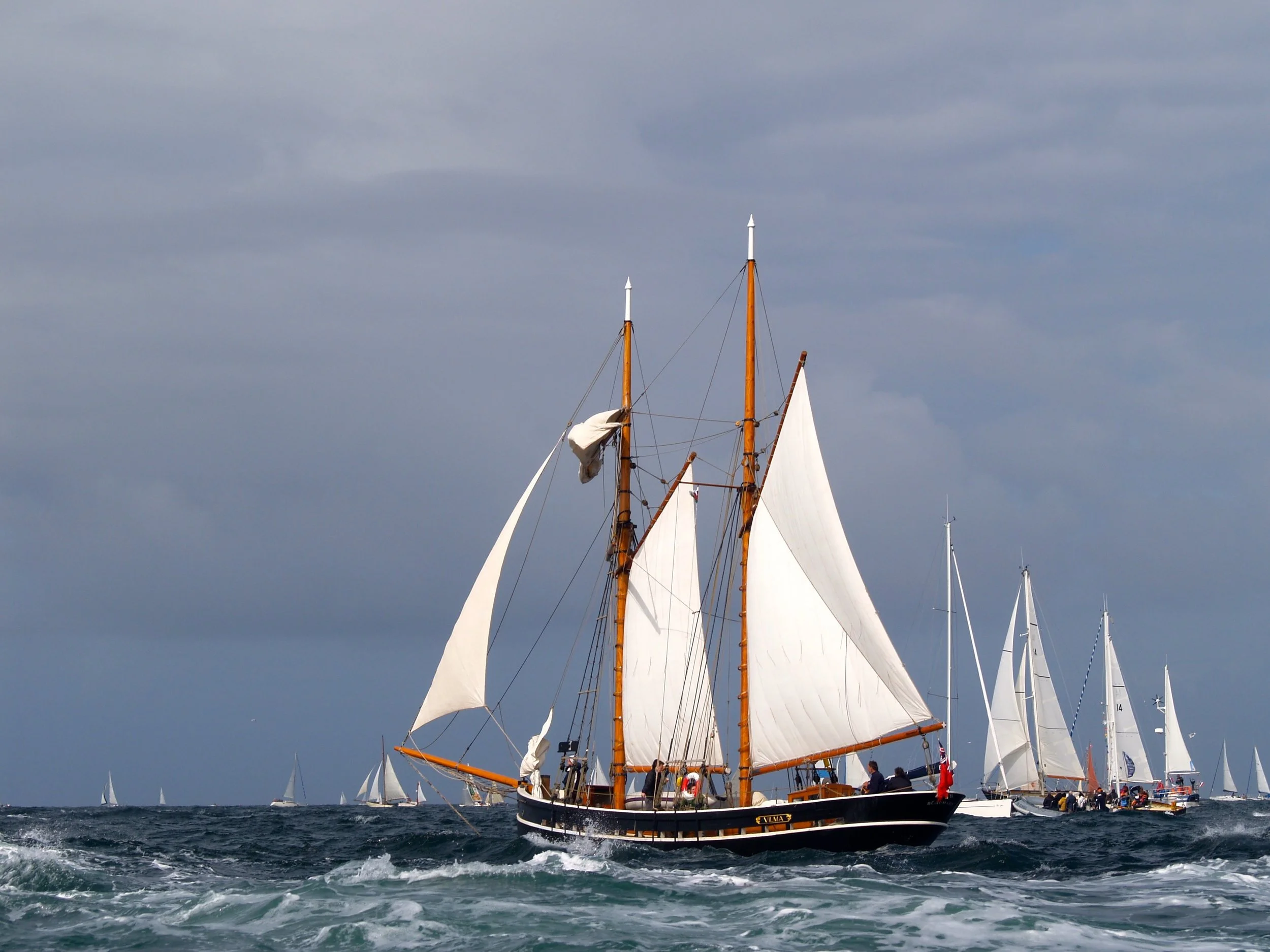 Un grand voilier avec des voiles blanches et un mât en bois navigue sur la mer, avec un ciel nuageux en arrière-plan, ainsi que d'autres petits voiliers en arrière-plan.
