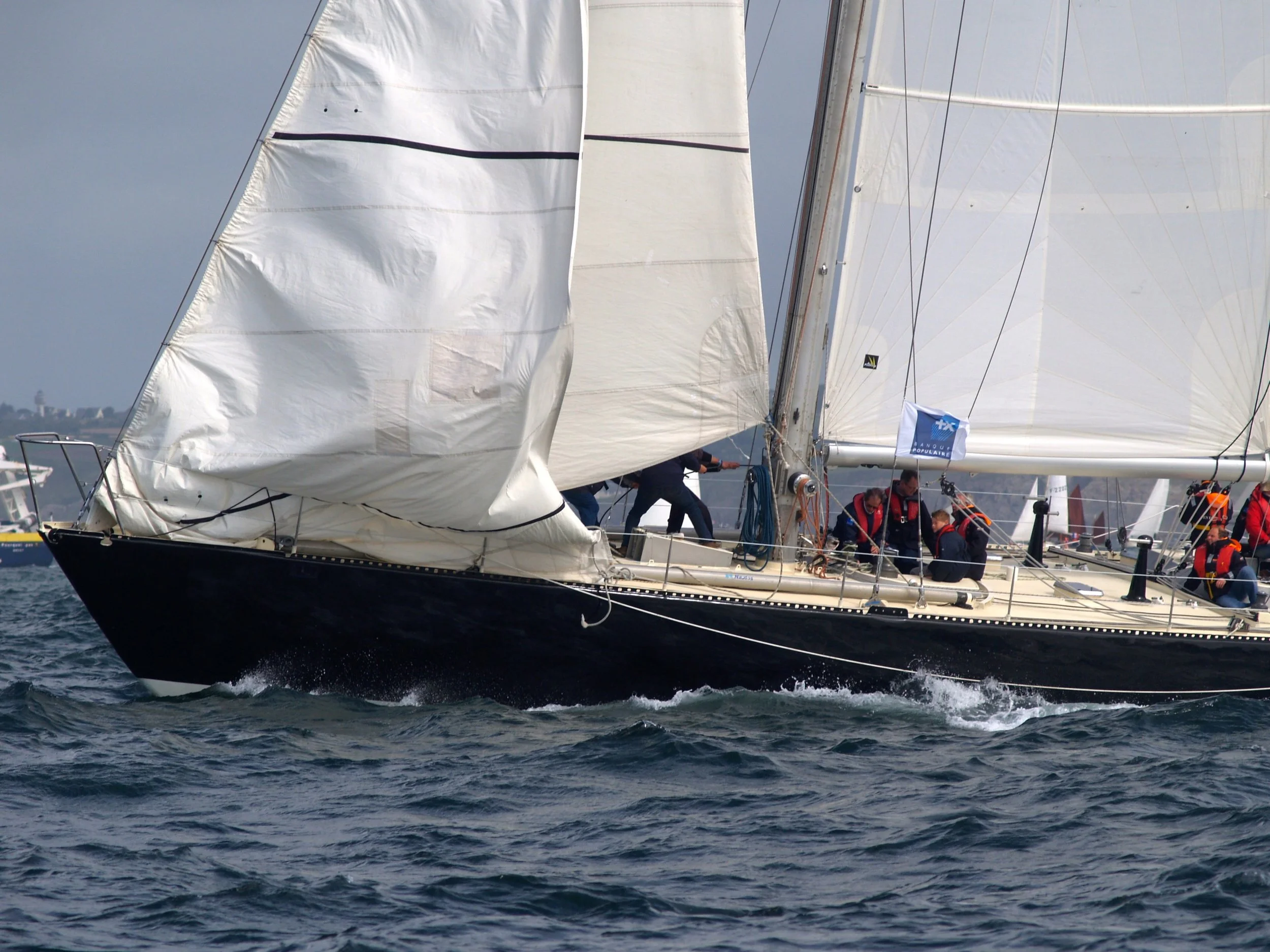 Un voilier en mer avec plusieurs personnes à bord, les voiles gonflées par le vent, lors d'une course ou d'une navigation à grande vitesse.