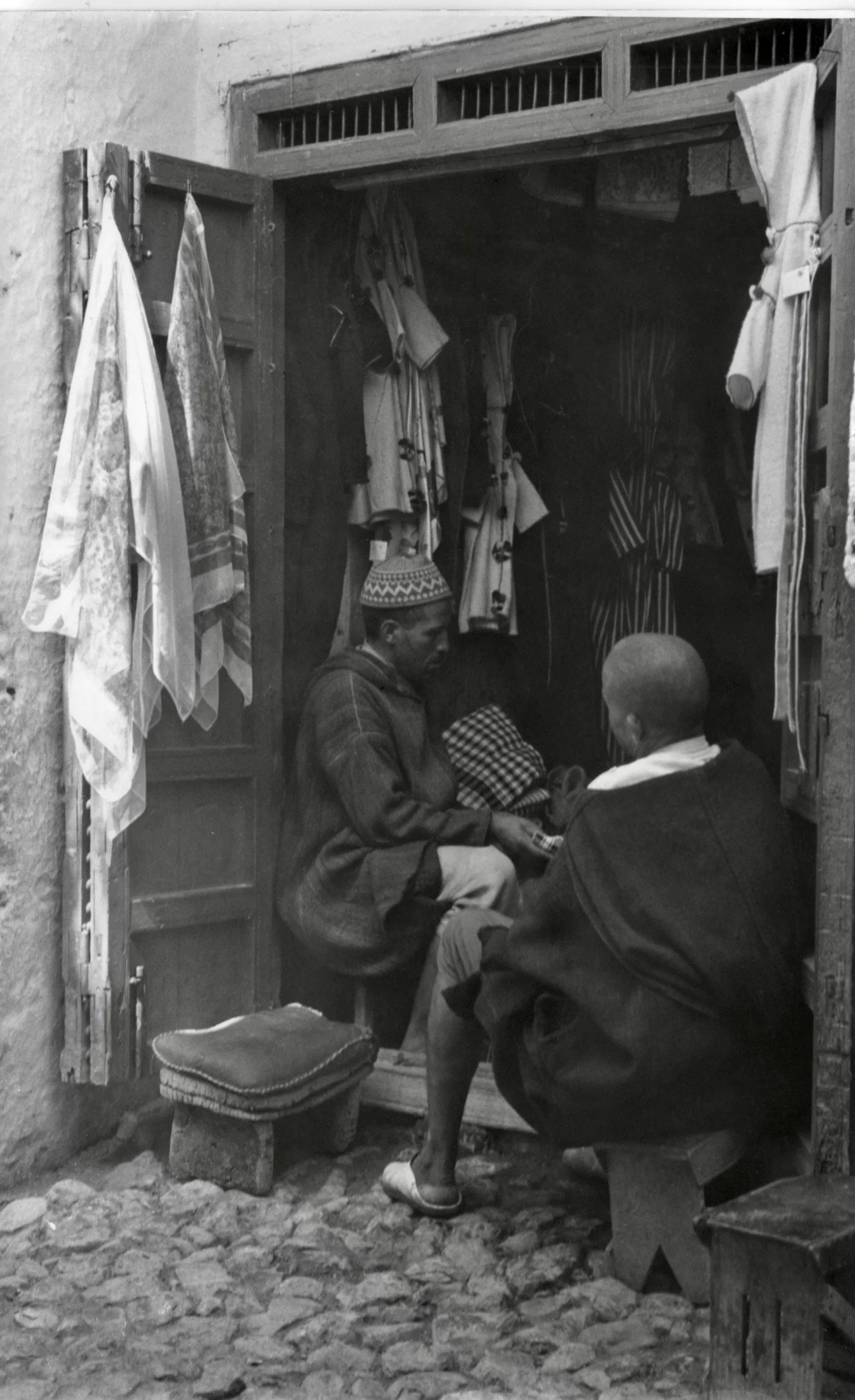 Deux hommes assis et une jeune fille en robe à motifs assise devant un salon de thé de rue avec des vêtements suspendus au dessus de l'entrée.