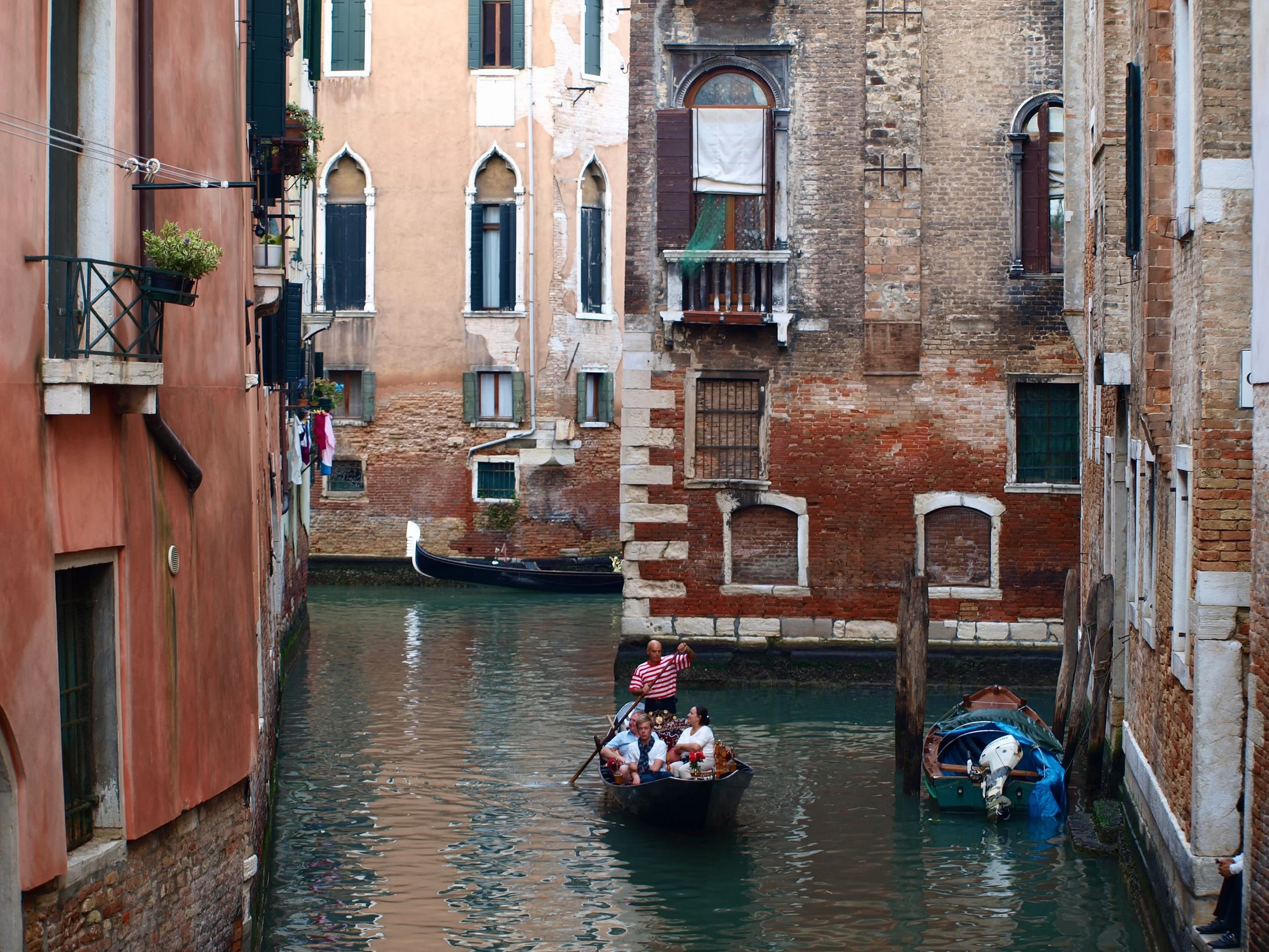 Une gondole navigue dans un canal étroit à Venise, entourée de bâtiments anciens en brique avec fenêtres à volets et balcons. Un guide en marinière dirige la gondole avec des touristes à bord. Le canal est calme, avec deux autres petites embarcations