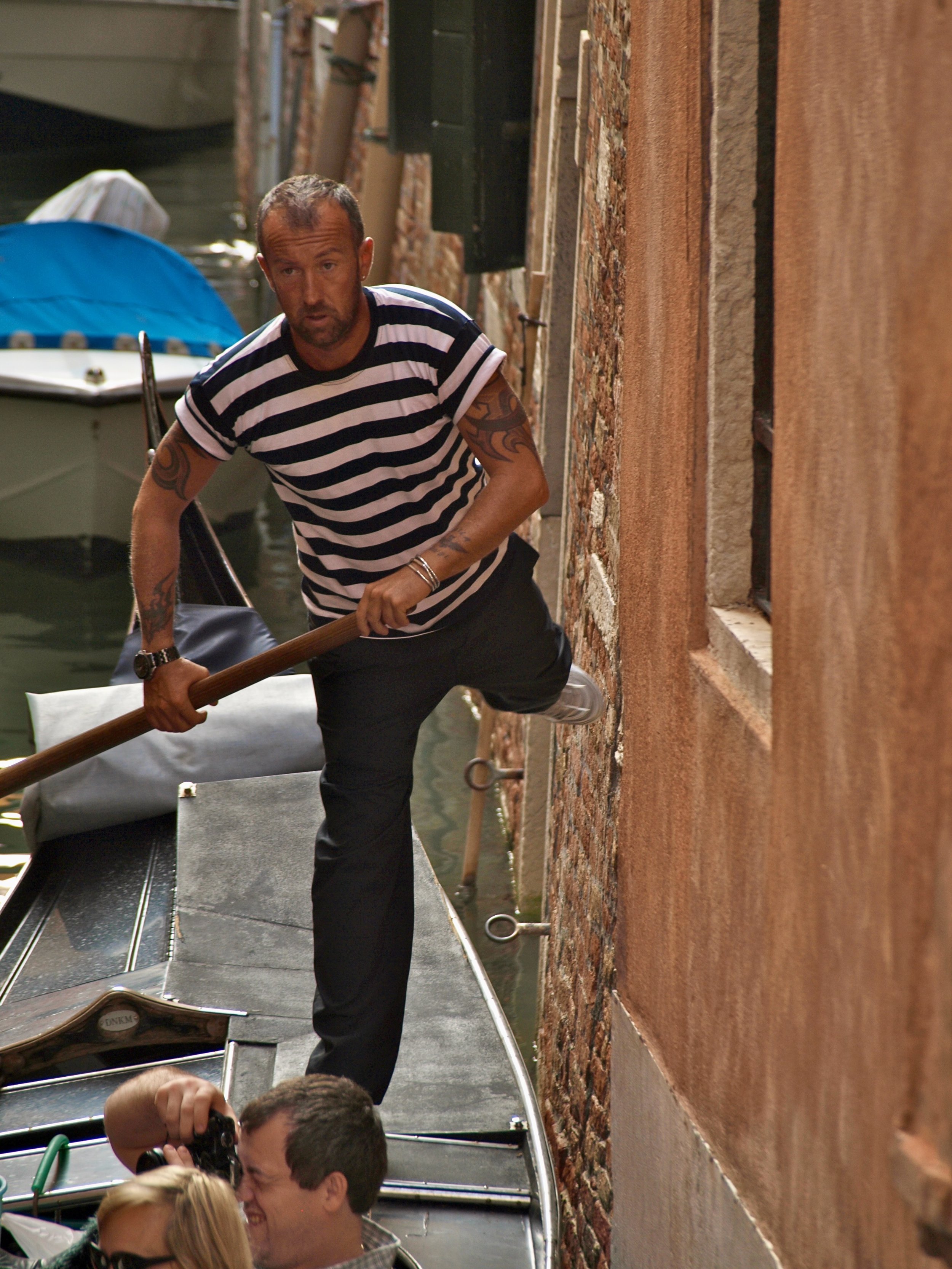 Un homme en maillot à rayures noires et blanches pousse une barque sur un canal étroit, avec des personnes à bord et des bâtiments en pierre rouge à côté.
