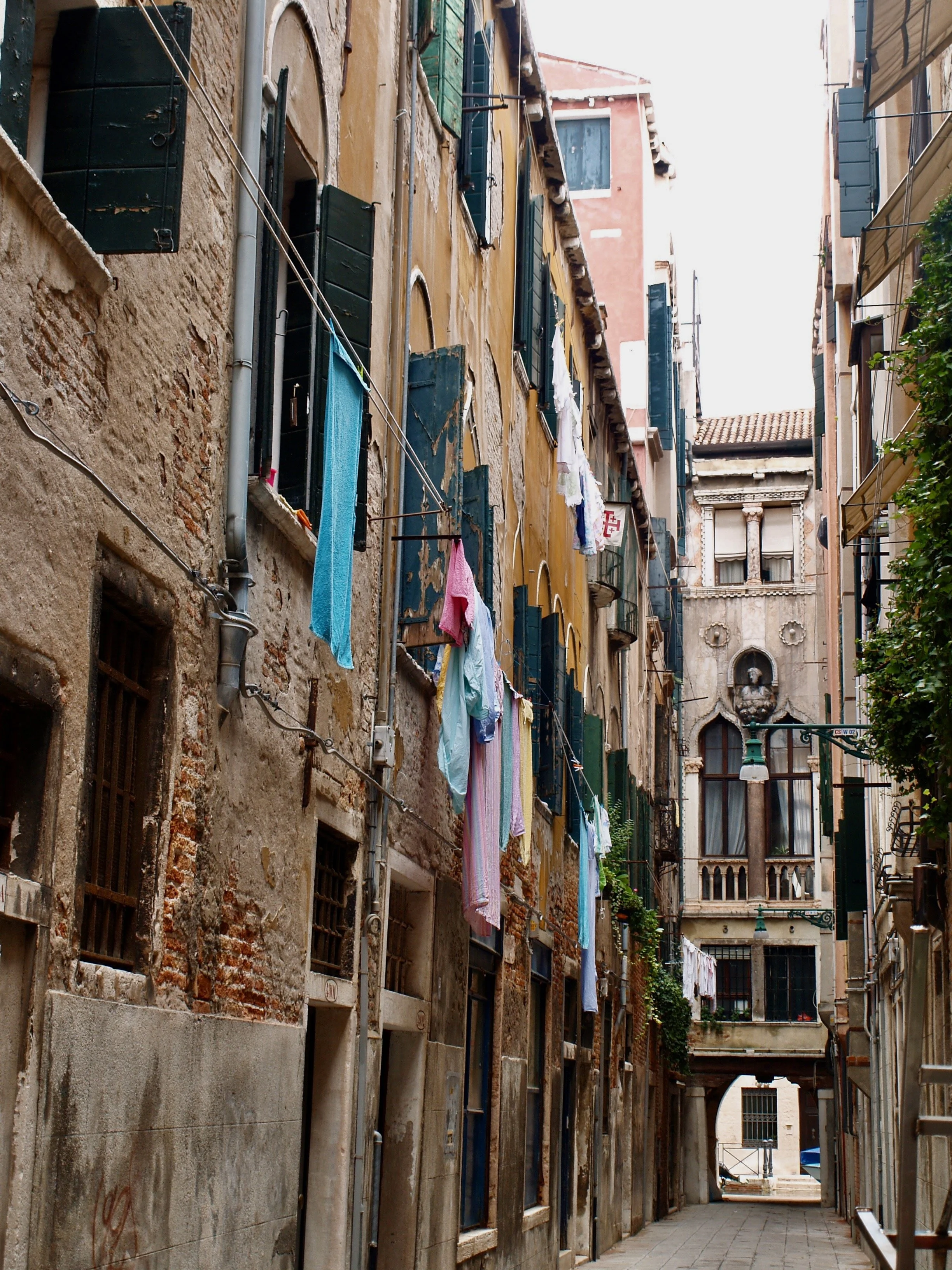 Vue d'une ruelle étroite avec des bâtiments anciens en pierre, certains avec des volets verts, des vêtements suspendus pour sécher en ligne, et une arche à l'extrémité de la ruelle.