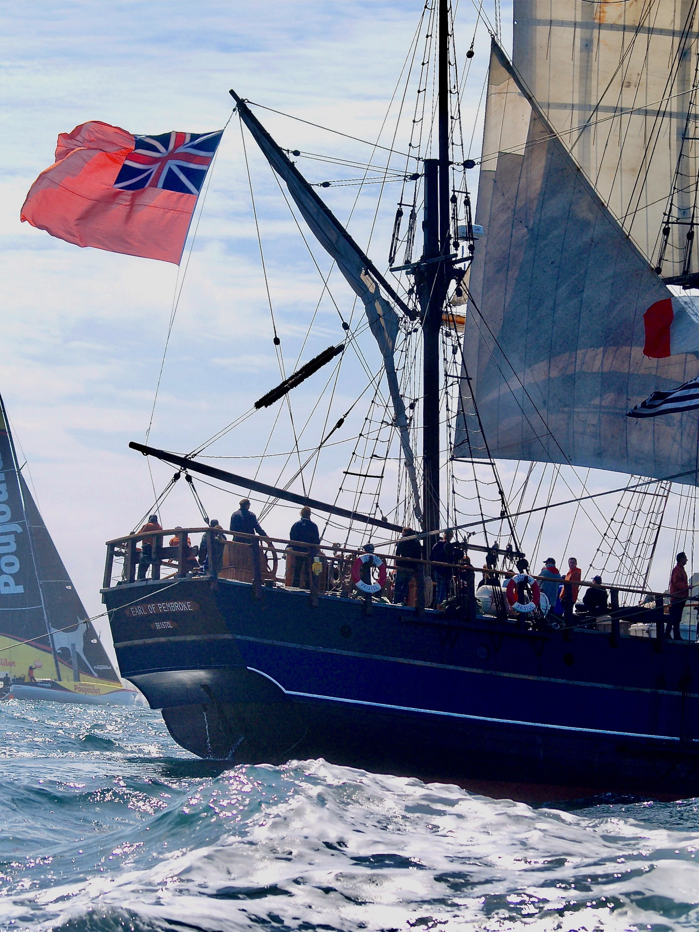 Un grand voilier avec un drapeau britannique en haut de sa mâture, naviguant en mer avec des personnes à bord. Le voilier porte le nom 'Earl of Pembroke' et la zone environnante montre des vagues et une ambiance ensoleillée.