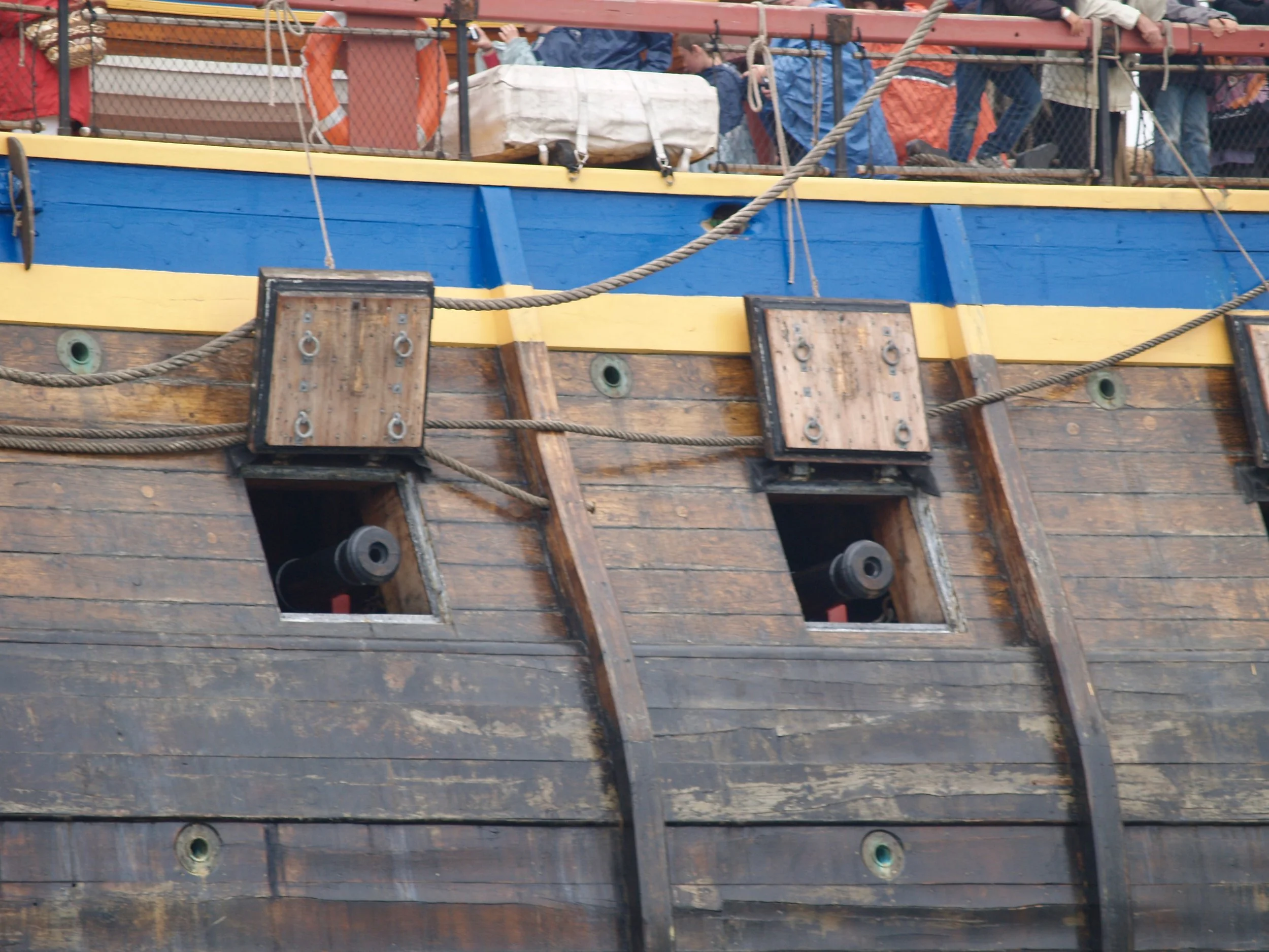 Un bateau en bois avec des cordages, des planches et des détails en couleur bleue et jaune, vue partielle.