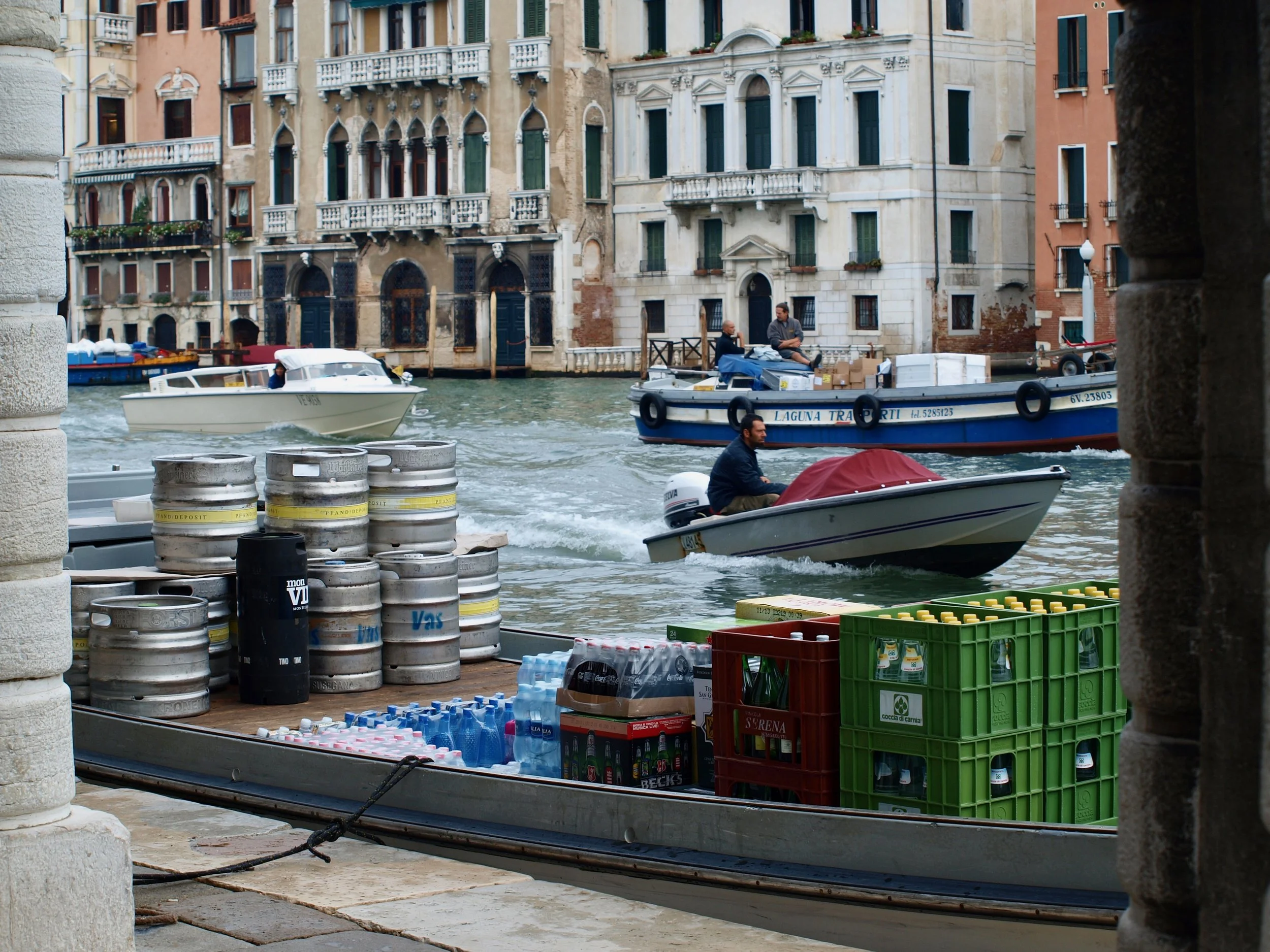Vue d'un canal à Venise avec des bâtiments anciens en arrière-plan, des bateaux naviguant sur l'eau, et des caisses contenant des bouteilles d'eau et de boissons, ainsi que des tonneaux de bière sur le quai.