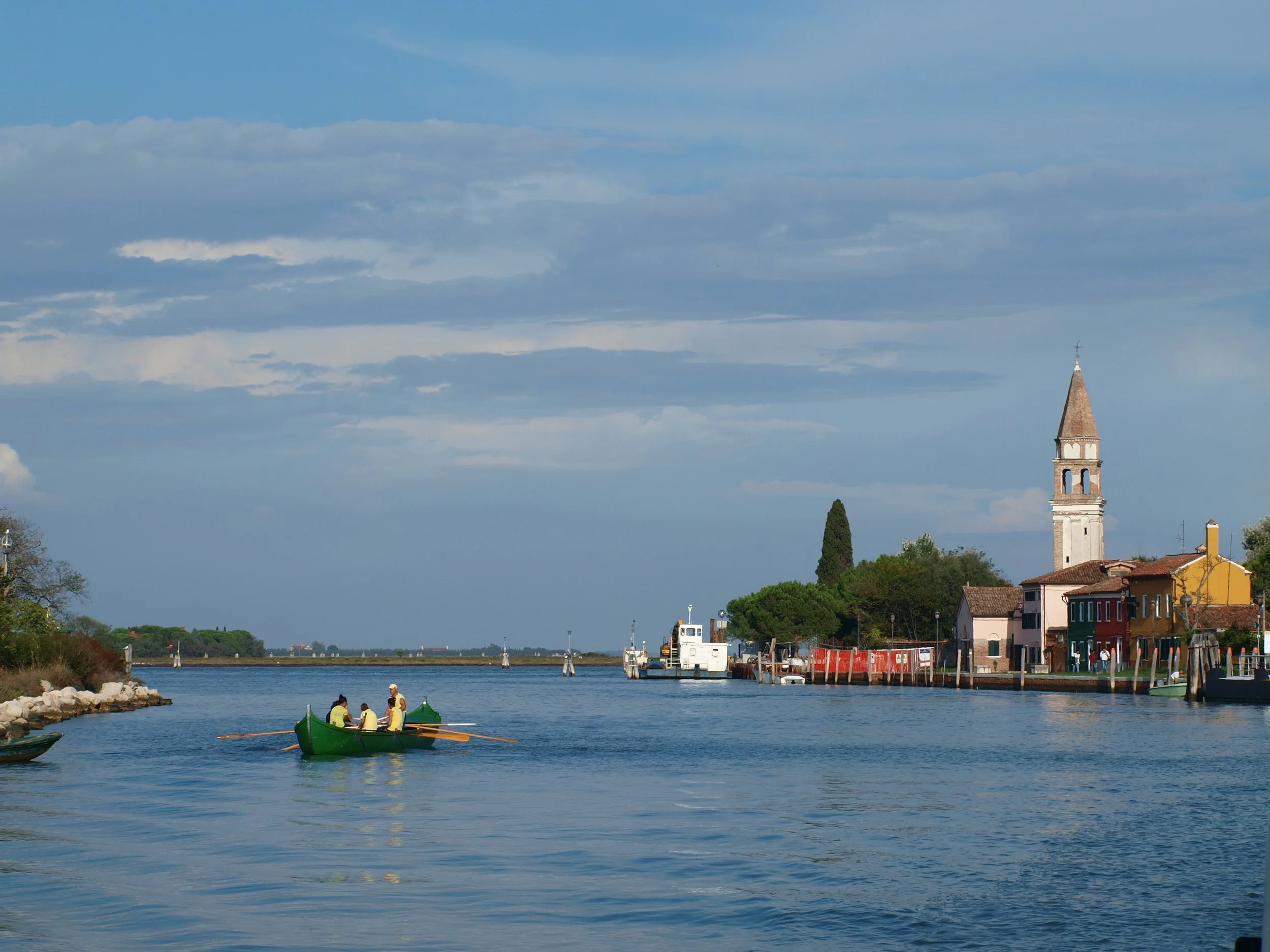 Scène avec une rivière, un bateau à rames avec quatre personnes, bâtiments colorés le long de la rivière, un clocher d'église, ciel partiellement nuageux.