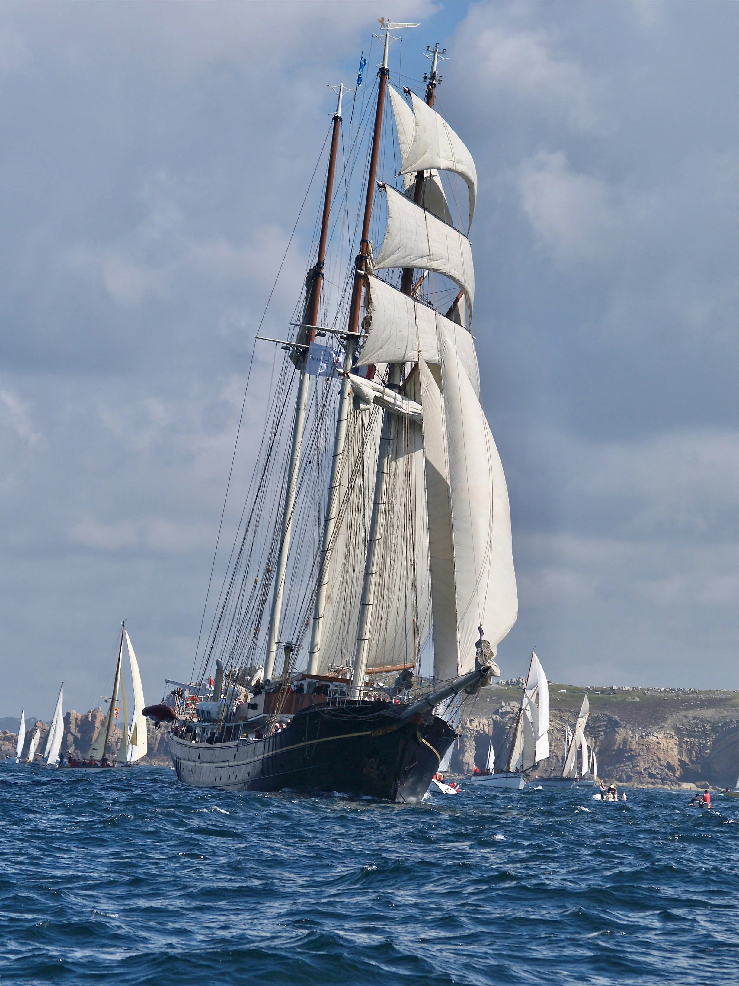 Un grand voilier avec des voiles blanches naviguant dans la mer, avec d'autres petits voiliers et une côte rocheuse en arrière-plan sous un ciel partiellement nuageux.