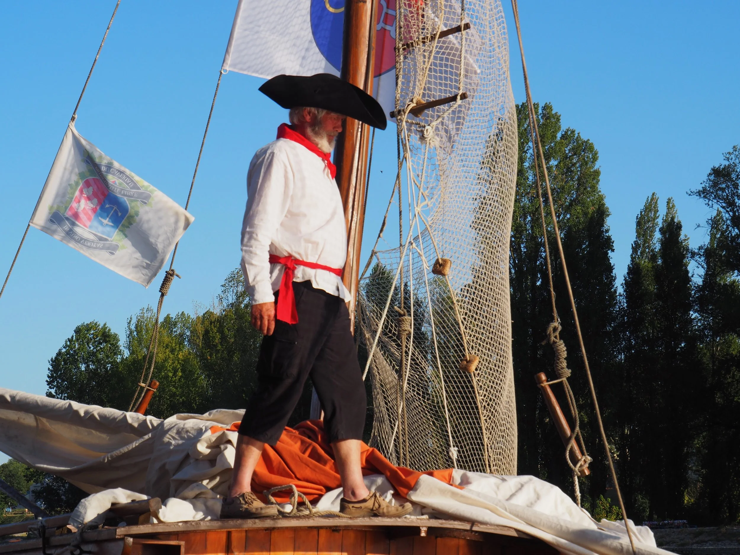 Un homme en costume colonial avec un chapeau noir, une chemise blanche, un foulard rouge, et une barbe blanche, se tenant à bord d'un bateau à voiles en bois, avec une voile et une grande grille en filet derrière lui, sous un ciel bleu clair, entouré