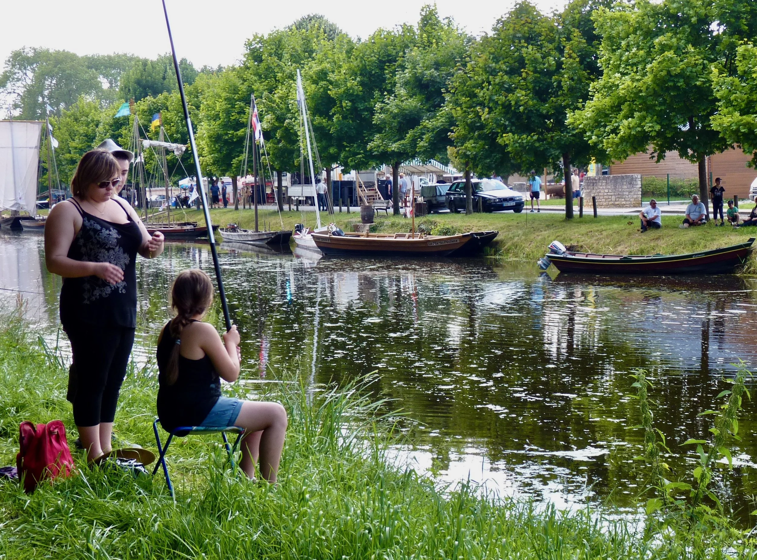 Jeune femme et enfant assis au bord d'une rivière, pêcheurs en arrière-plan, bateaux à voile amarrés, arbres verts, activité de loisirs en plein air.