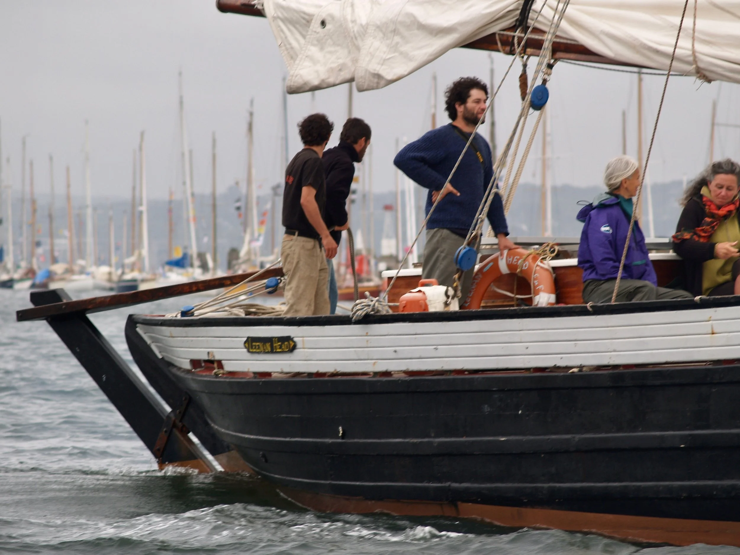 Groupe de personnes à bord d'un voilier, naviguant en mer par une journée nuageuse, avec d'autres voiliers visibles en arrière-plan.