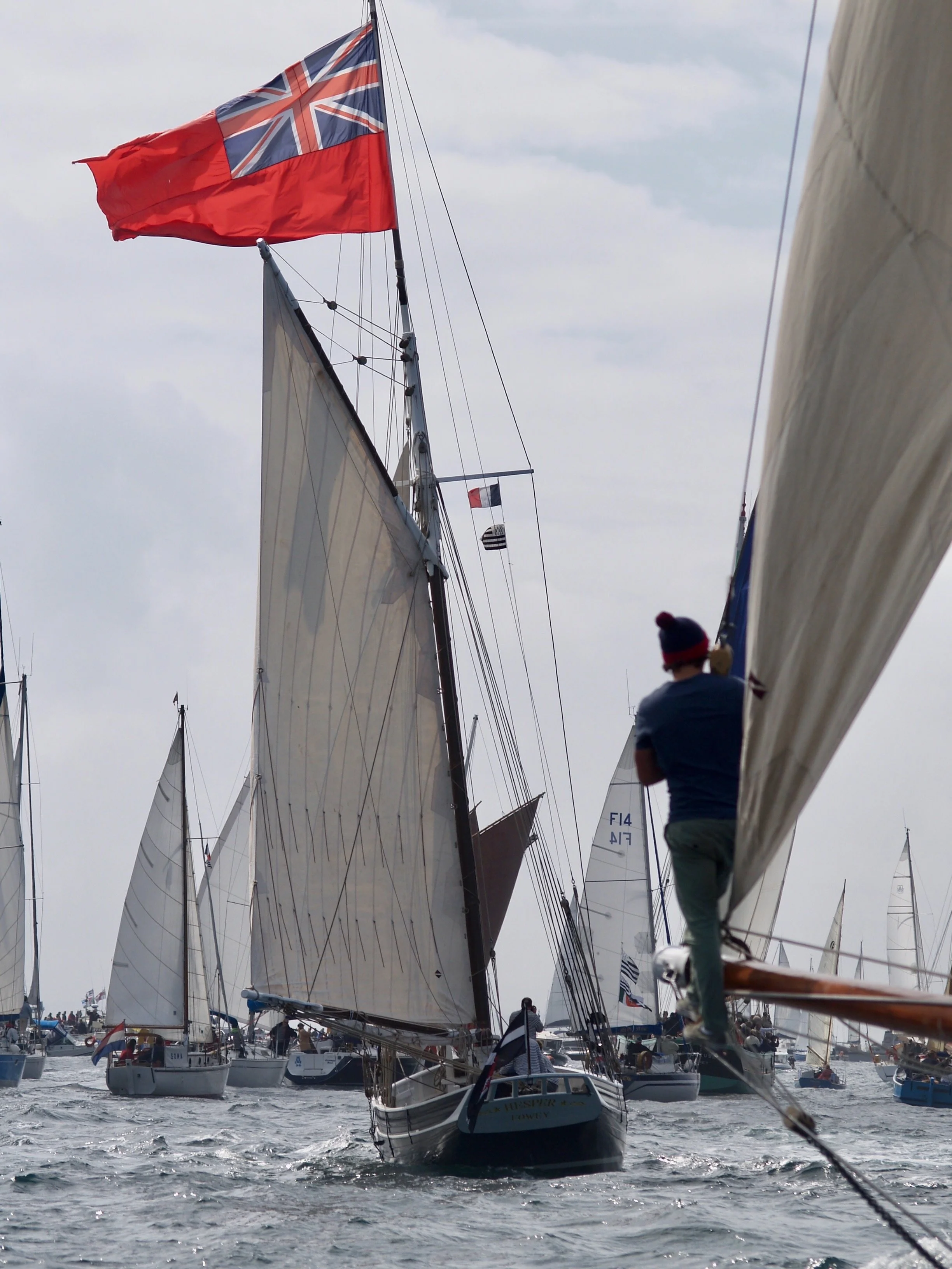 Des voiliers en mer lors d'une régate, avec un drapeau britannique en haut et des voiles blancs, un homme en bleu et chapeau rouge sur un bateau.