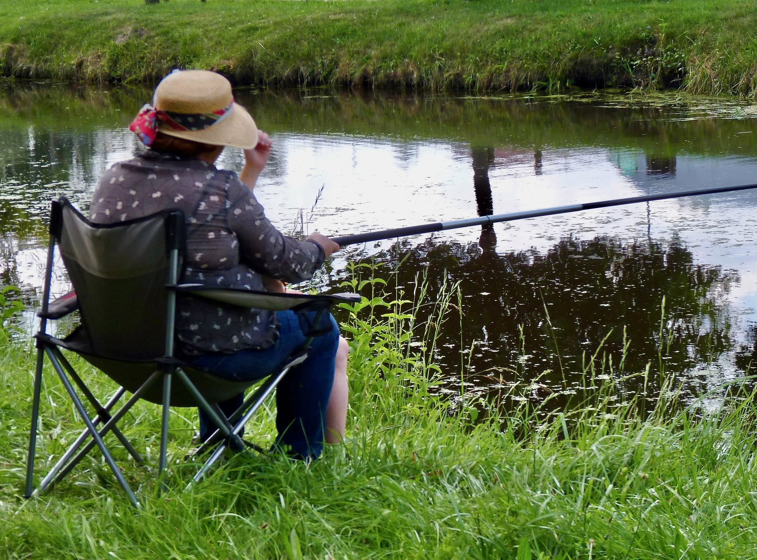 Une personne assise en fauteuil pliant regarde un étang ou une rivière en train de pêcher, entourée d'herbe verte et de végétation.