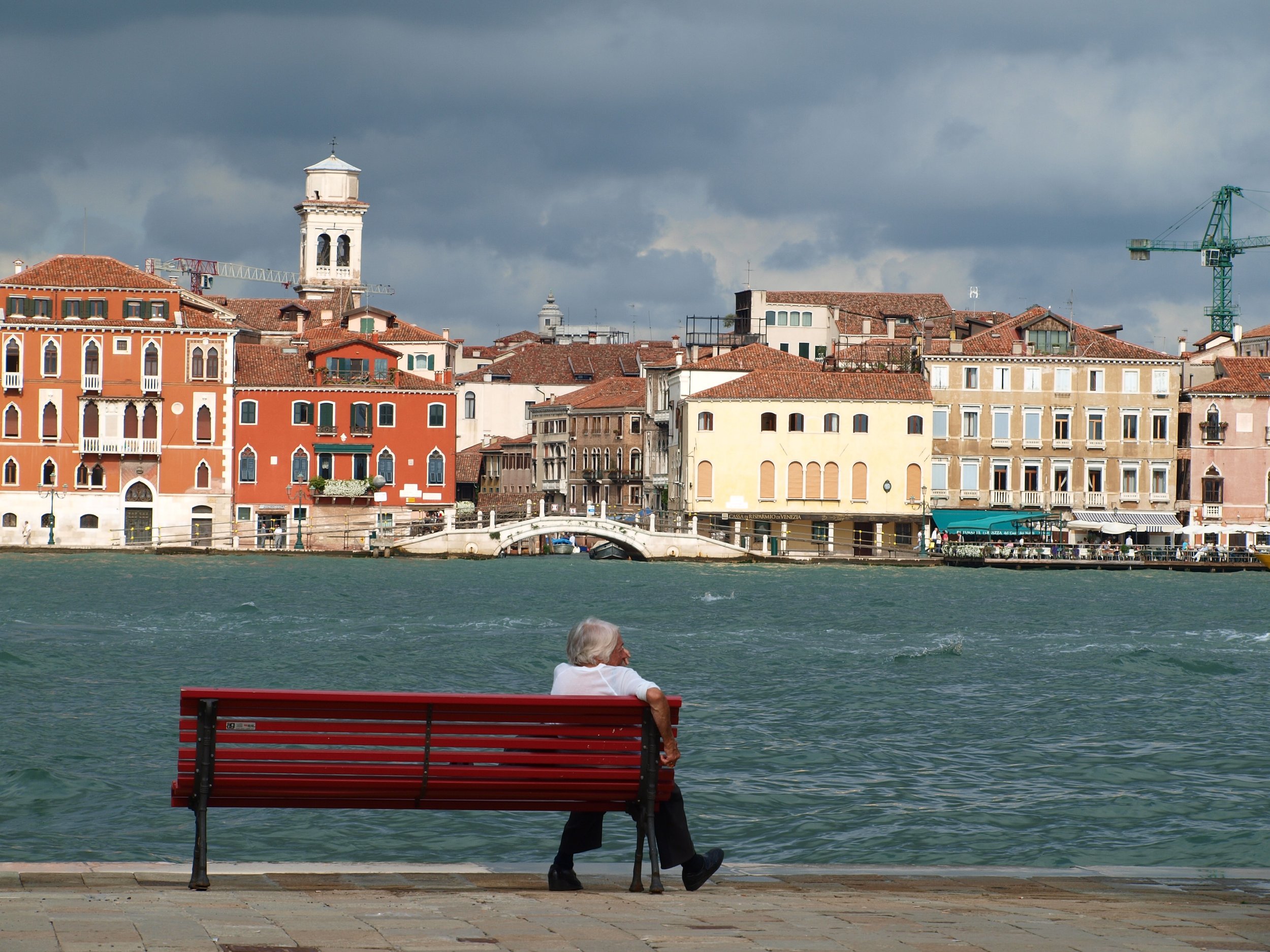 Une personne âgée assise sur un banc rouge près de l'eau, regardant la ville de Venise avec des bâtiments colorés et un ciel nuageux en arrière-plan.