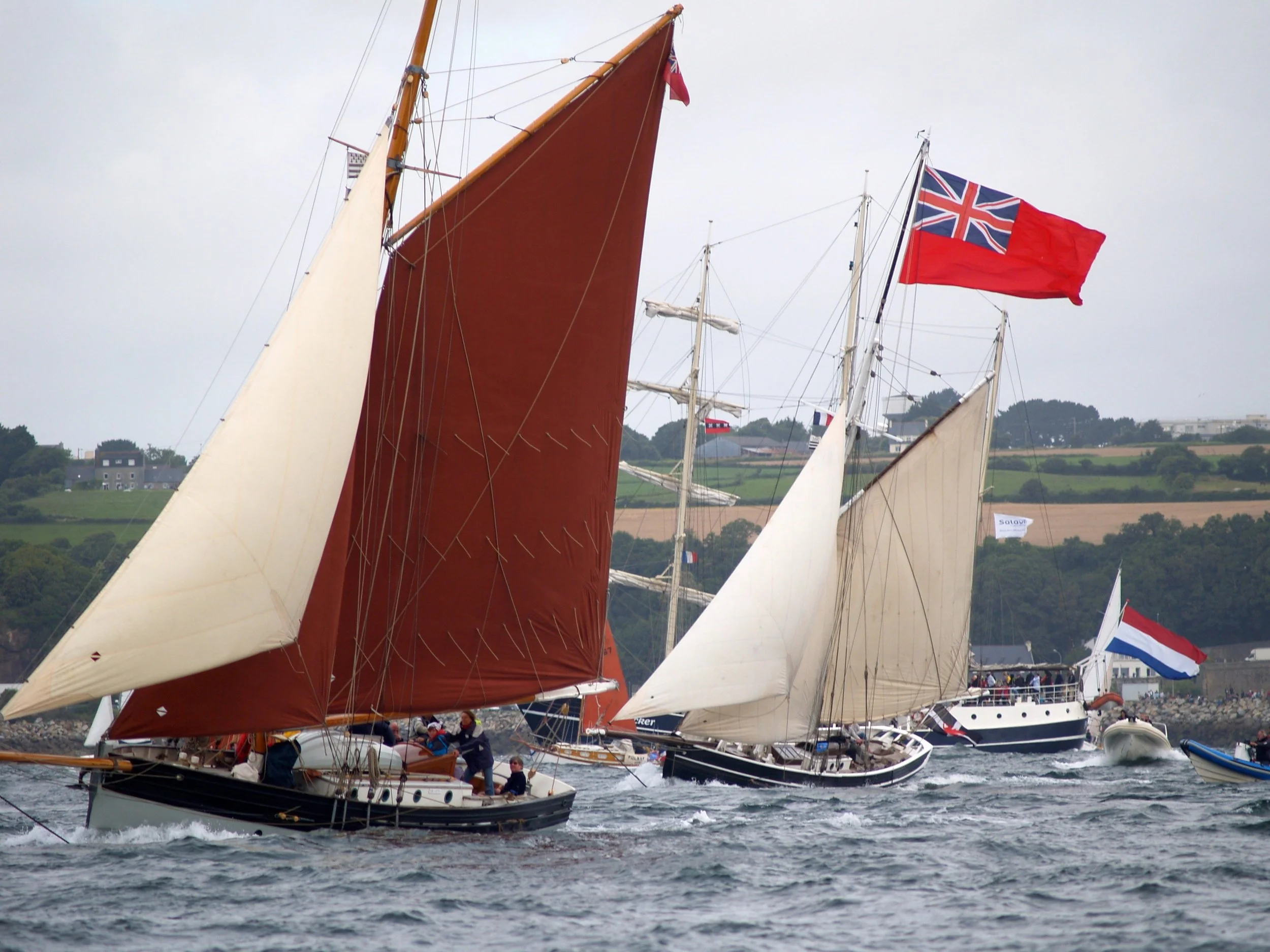 Des bateaux à voile avec des voiles rouges, blancs et à motifs de drapeaux britanniques et français, naviguant dans un port, avec des collines et des bâtiments en arrière-plan.
