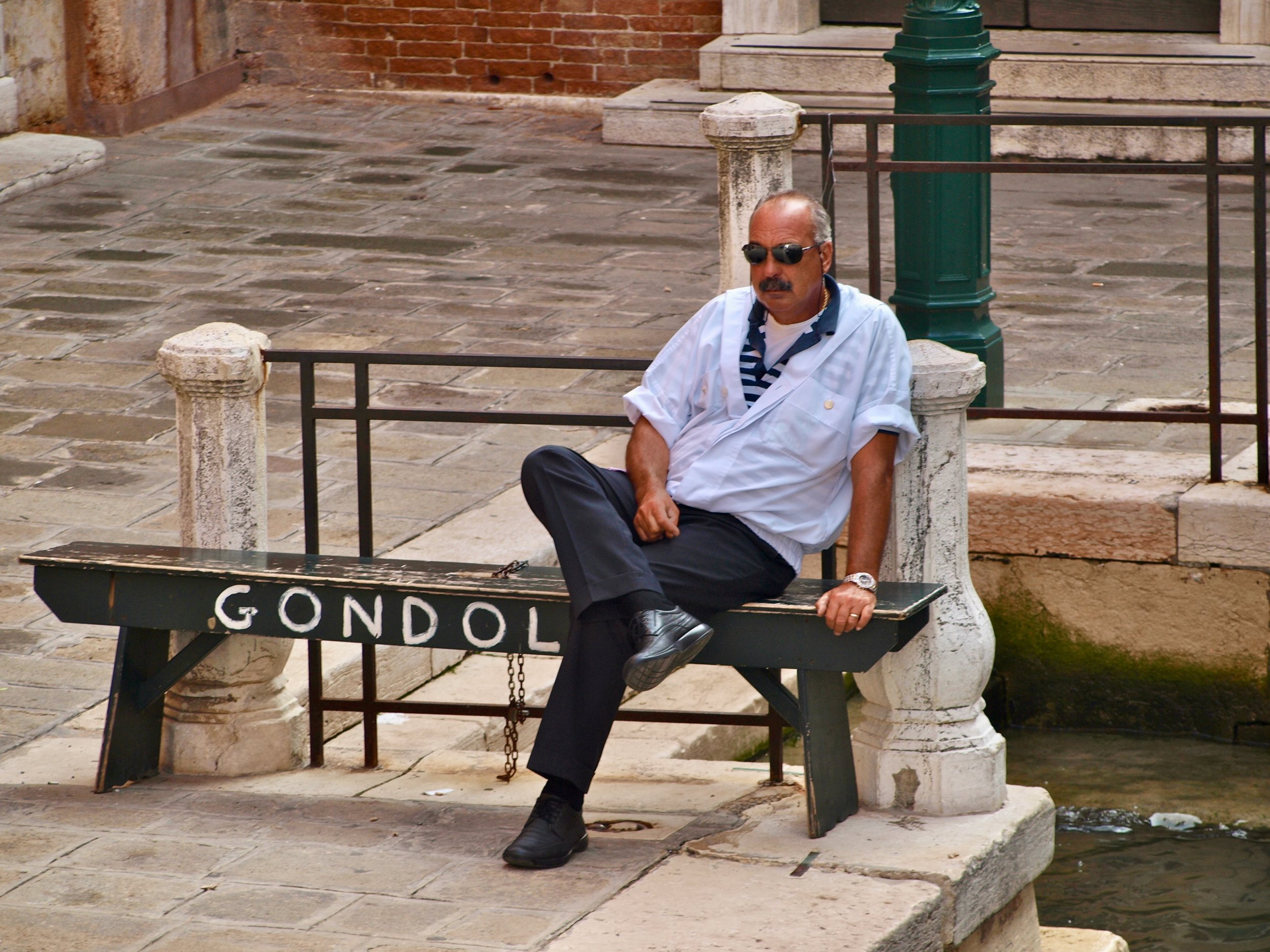 Un homme avec une moustache, portant des lunettes de soleil, une chemise blanche et un t-shirt à rayures, assis sur un banc. Il est à côté d'un rampe décorative en fer et d'une fontaine ou un monument avec une inscription en français. Le décor semble