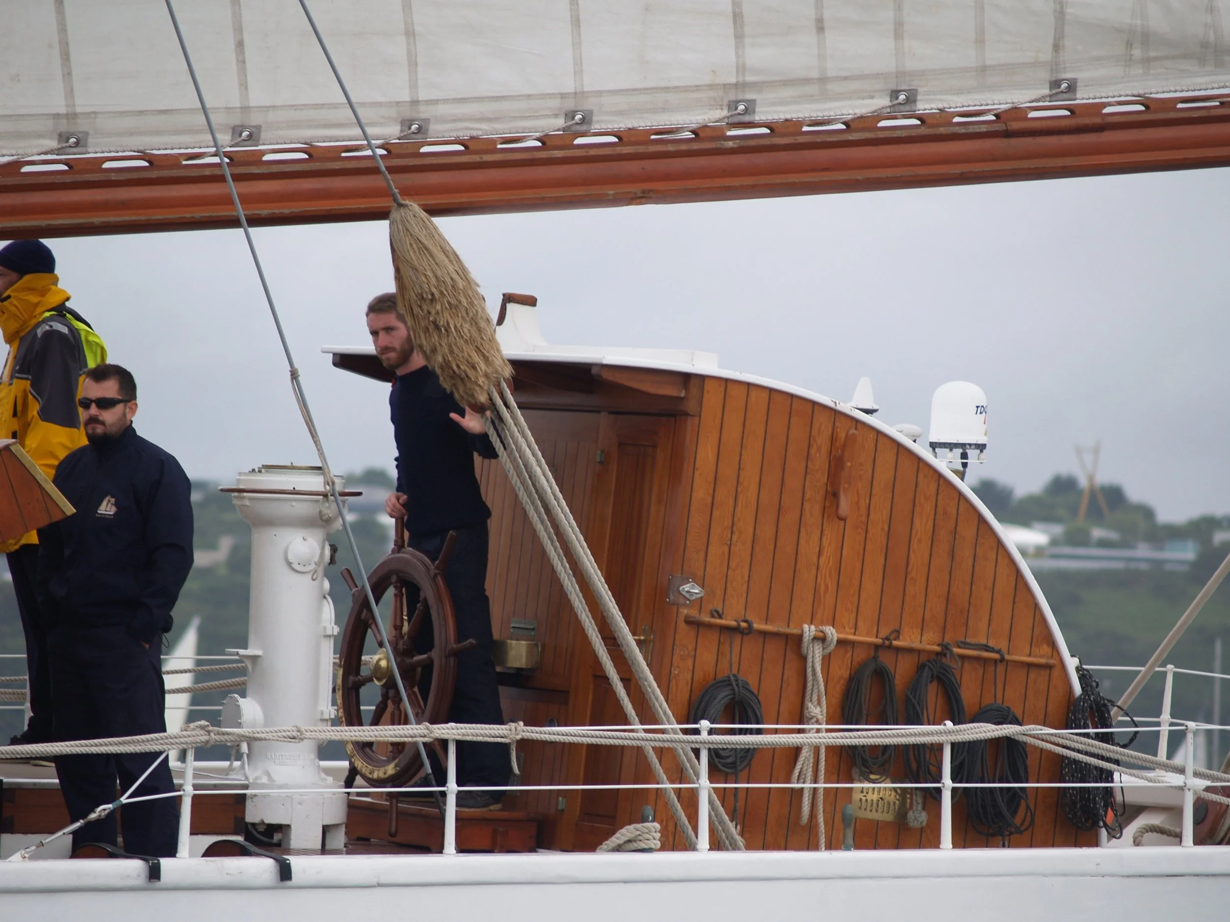 Trois hommes en vêtements de marin sur un bateau en bois. L'un d'eux tient une corde, tous portent des habits sombres et se trouvent sur le pont du bateau avec une boussole et des équipements marins visibles.